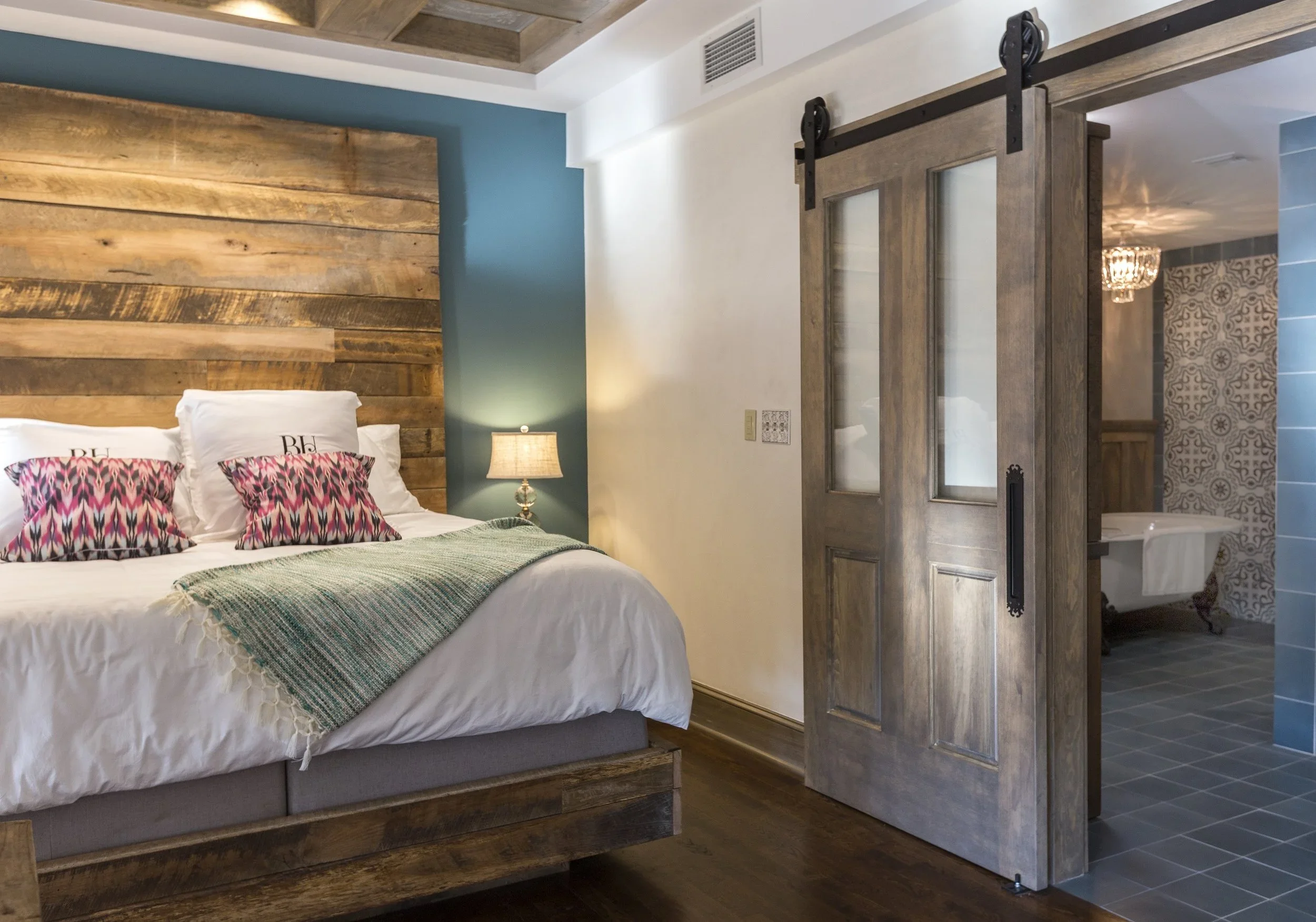 A cozy bedroom with a rustic wood headboard, white bedding, two decorative pillows, a nightstand with a lamp, and a sliding barn door leading to a bathroom with patterned tiles and a clawfoot tub.