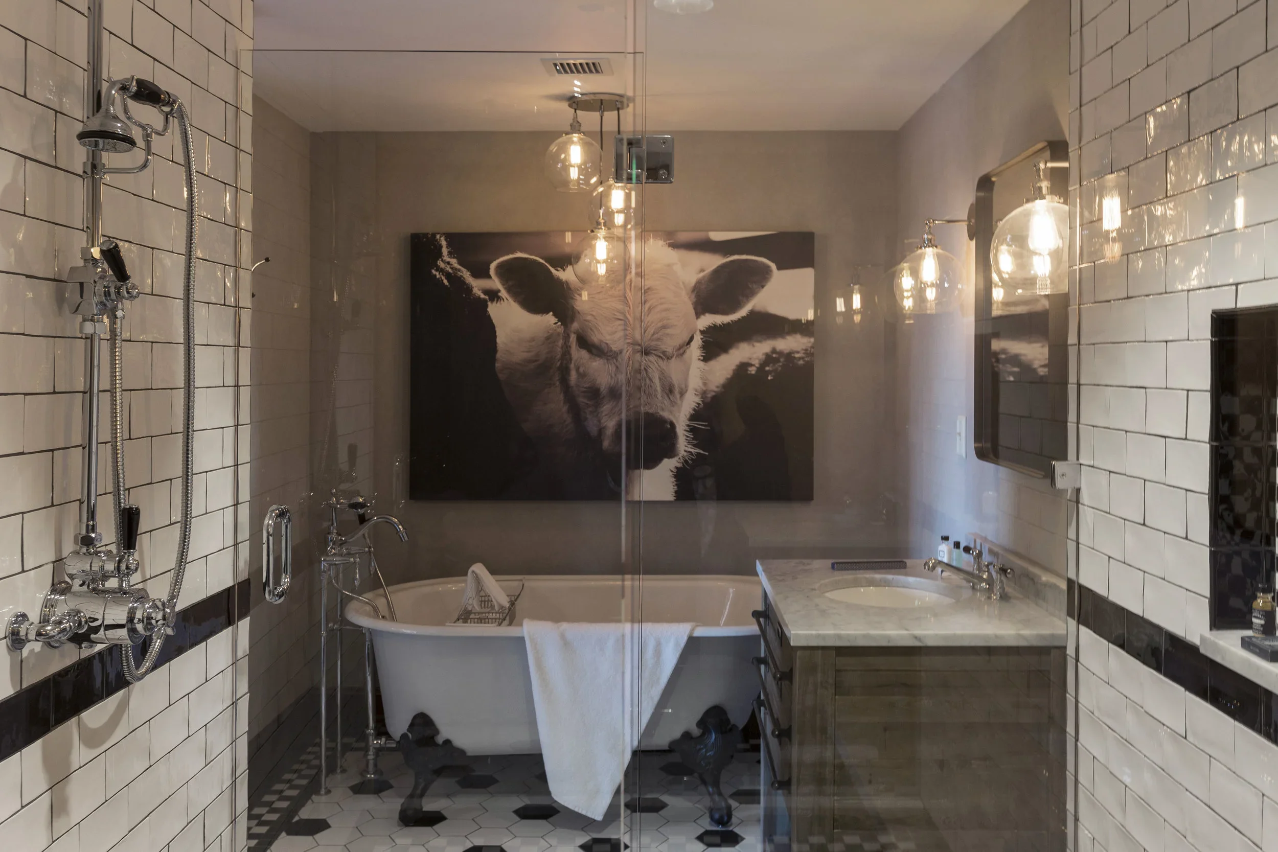 A bathroom with black and white tiled floor, a vintage clawfoot bathtub, a modern vanity with marble countertop, a large black and white cow portrait on the wall, and multiple hanging light bulbs.