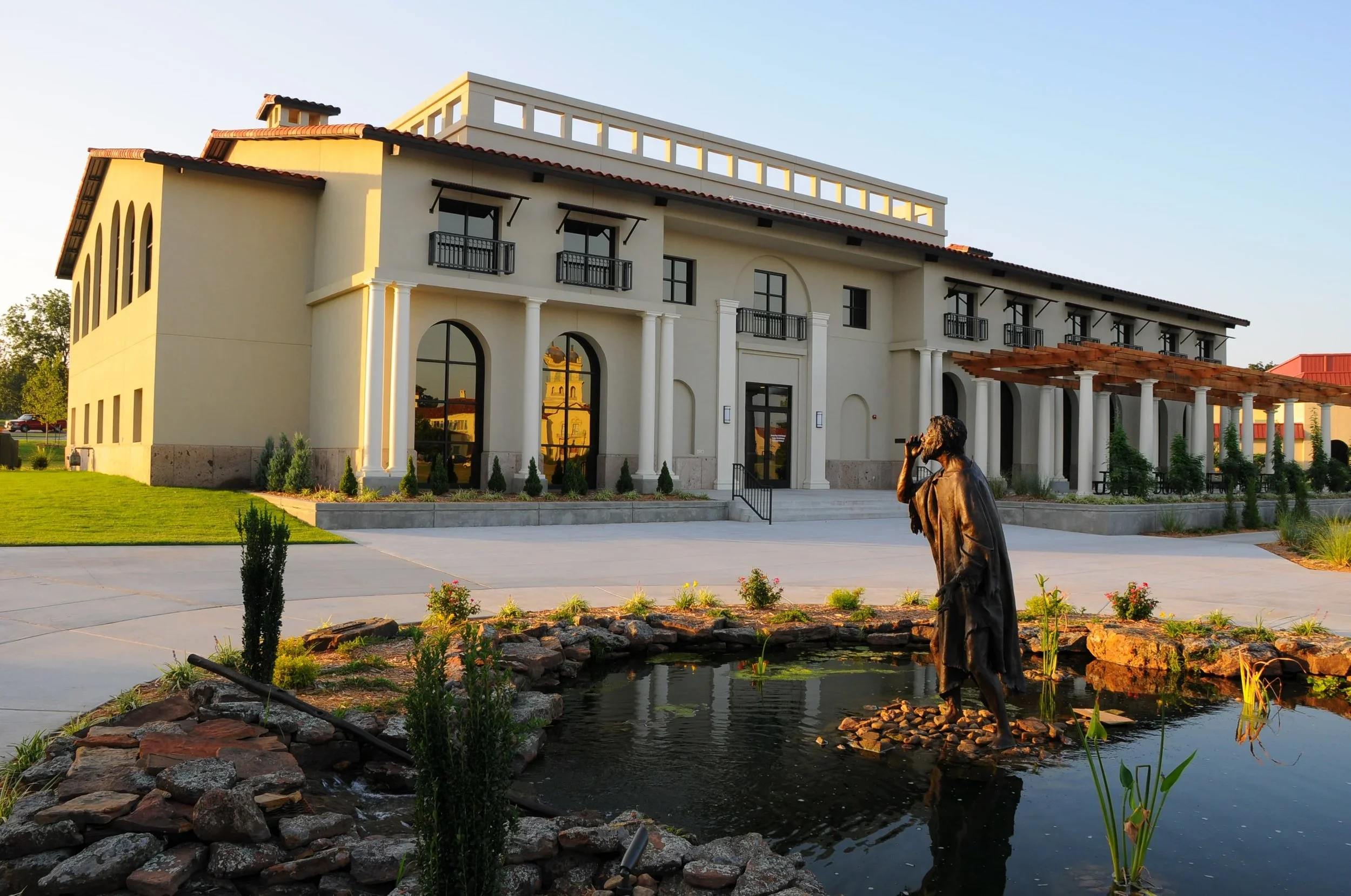 A large white building with arched windows, balconies, and a red tile roof, set near a pond with a statue of a person fishing, surrounded by landscaped greenery.