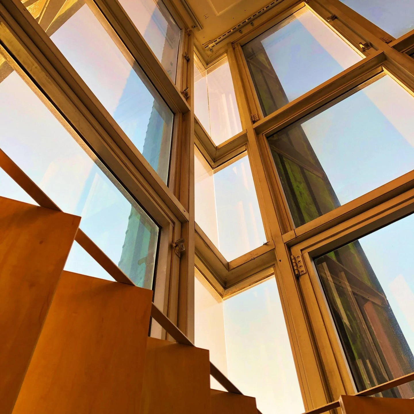View of wooden staircase from below looking up, with large floor-to-ceiling windows showing the sky outside.