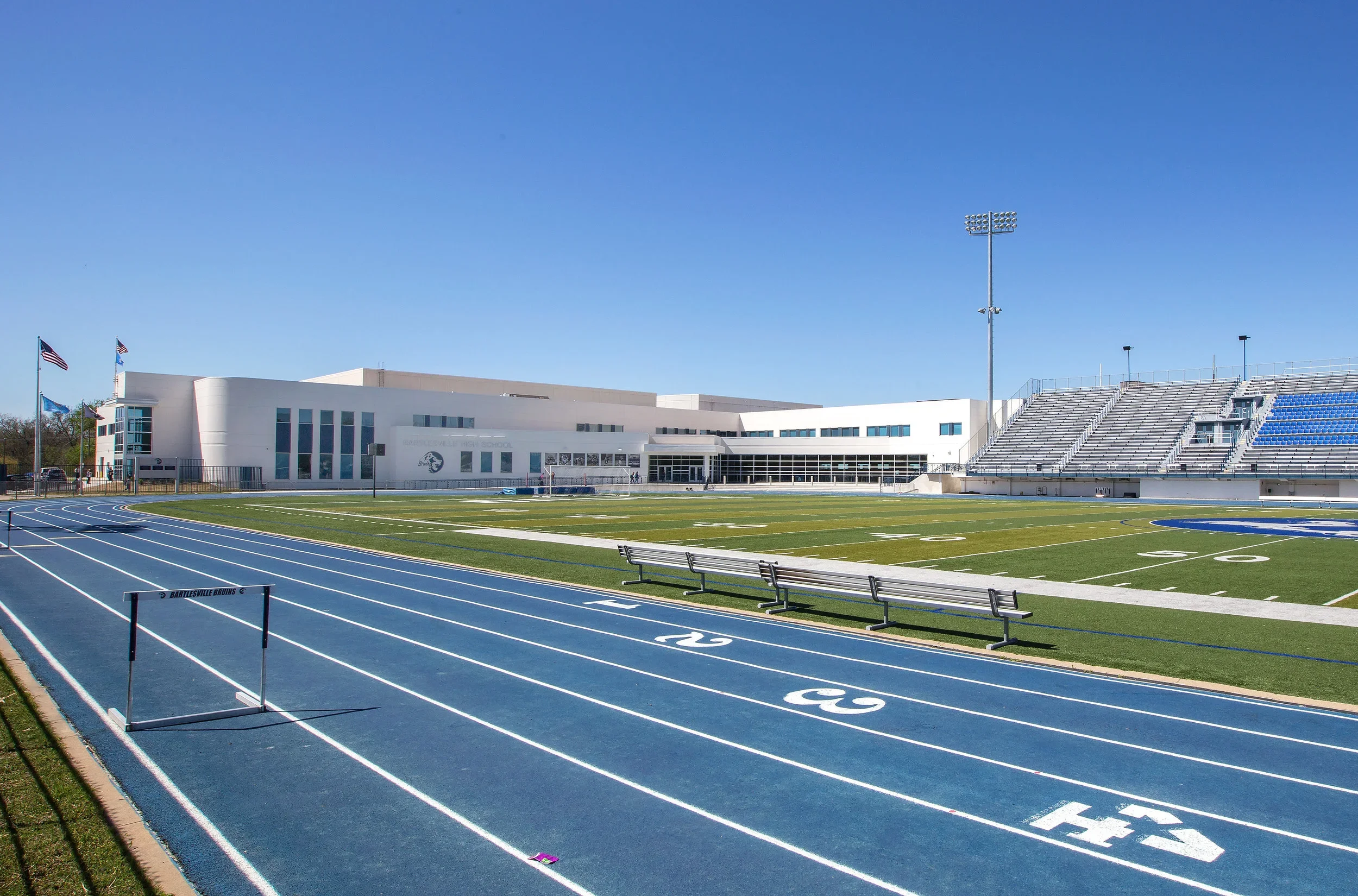 Outdoor sports stadium with a running track, football field, and bleachers. Clear blue sky, flags, and a modern school building in the background.