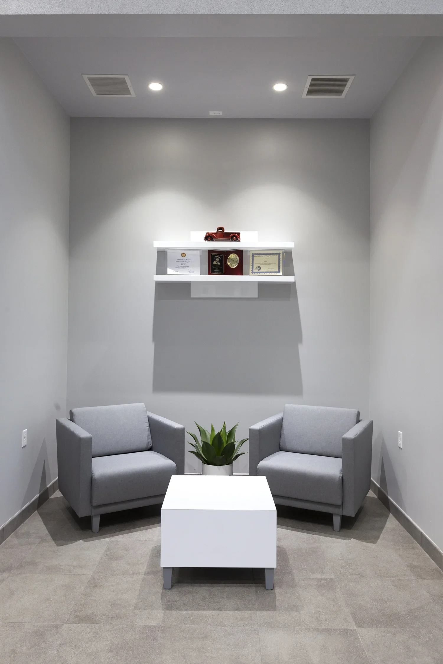A waiting area with two gray armchairs, a white coffee table, and a potted plant in the center. On the wall, a white shelf displays a toy truck, awards, and a framed certificate, with two ceiling lights above.