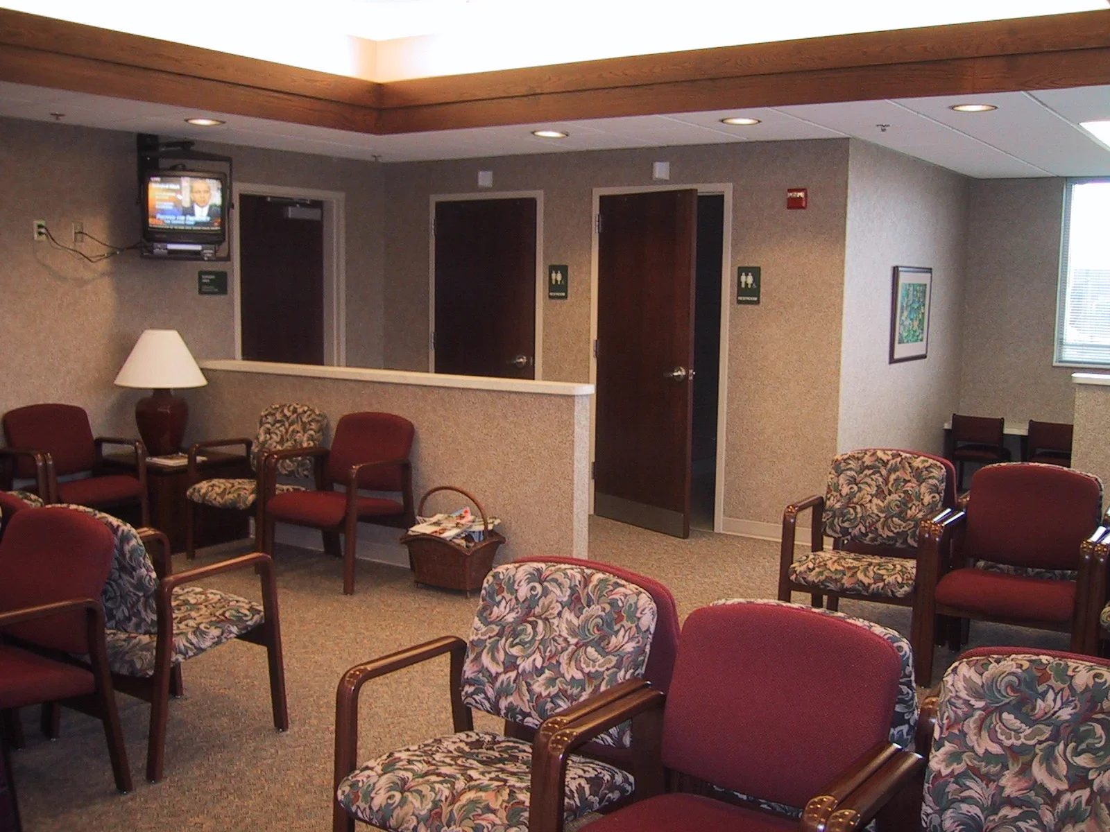 Waiting room with multiple chairs, a lamp, a small table, doors for restrooms, and a mounted television.