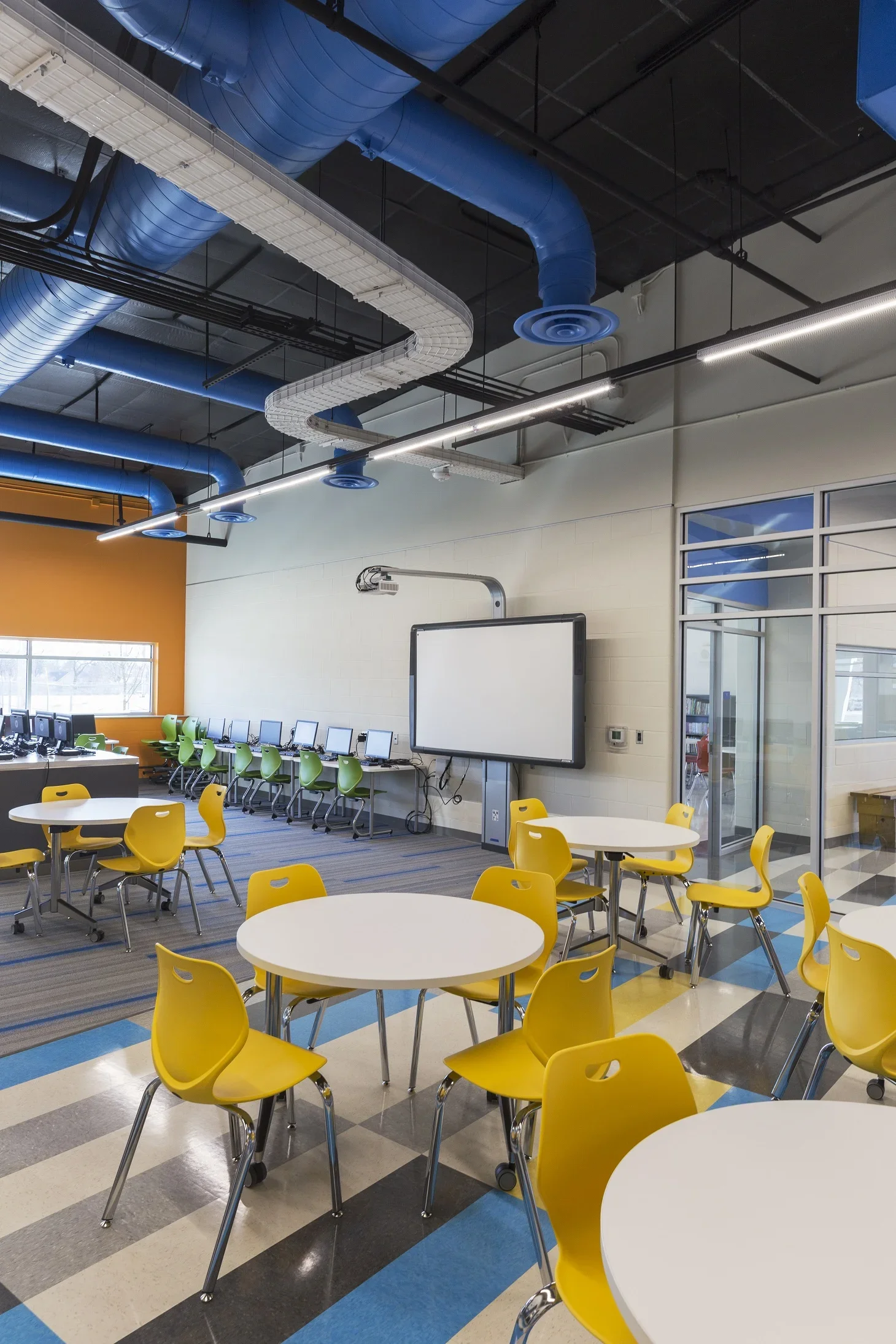 A modern classroom or computer lab with round yellow chairs, tables, and a row of computers along one wall. The room has a colorful, patterned floor, large windows, and an interactive whiteboard or smartboard on the wall. Exposed blue and white venti