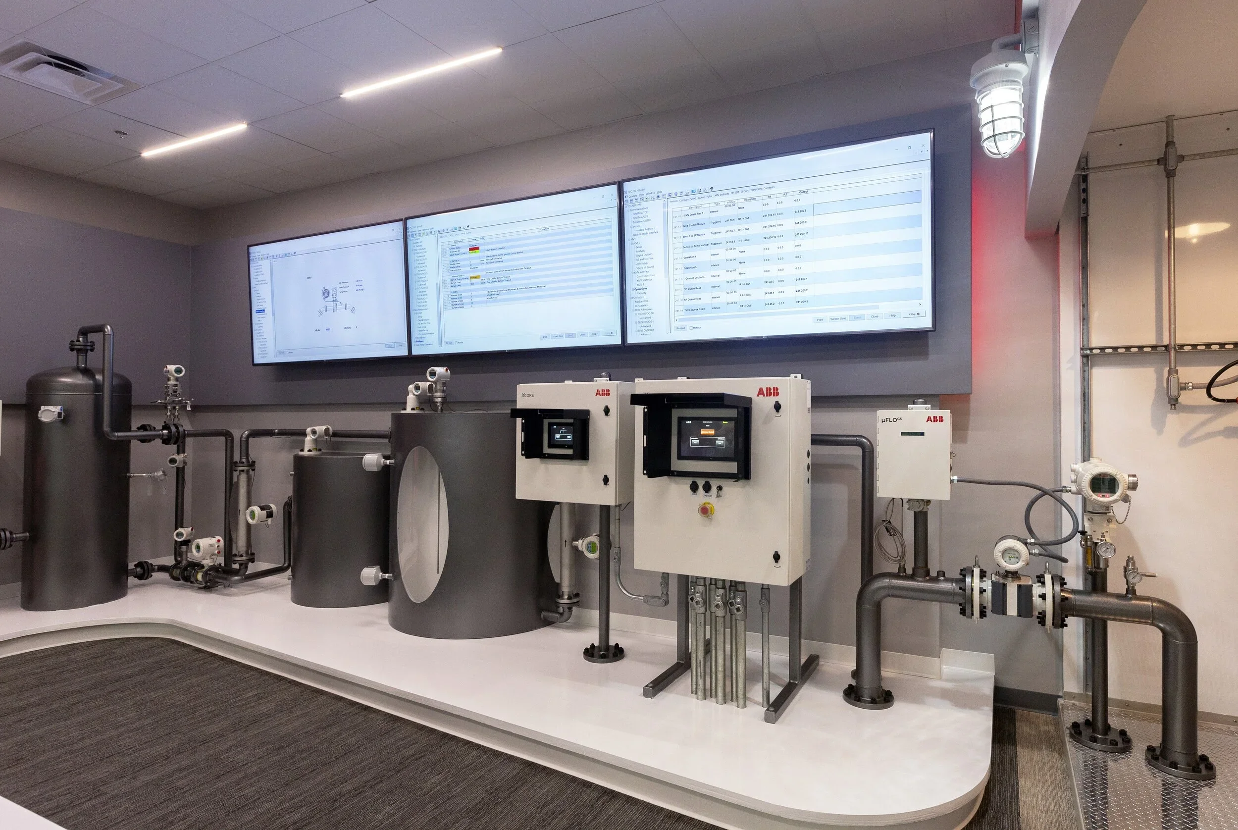 Industrial control room with multiple monitors displaying system data, and machinery with pipes and electronic panels.