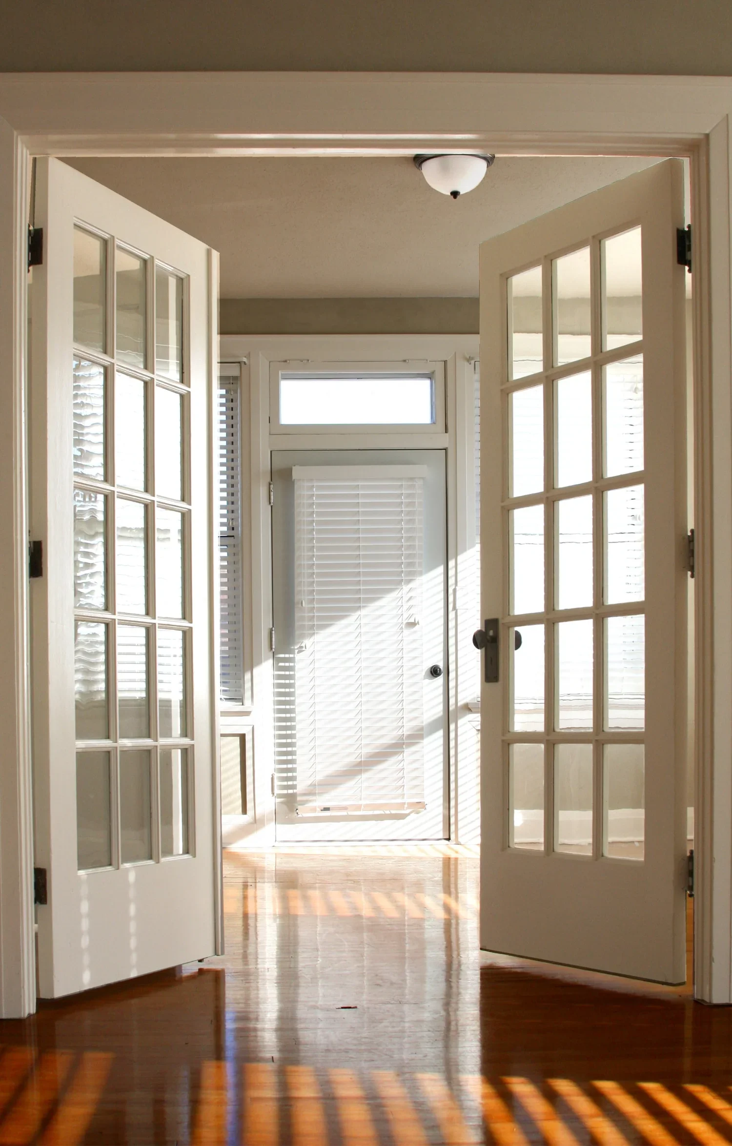 Open French doors leading into a bright room with a door and windows with blinds, sunlight casting shadows on the hardwood floor.