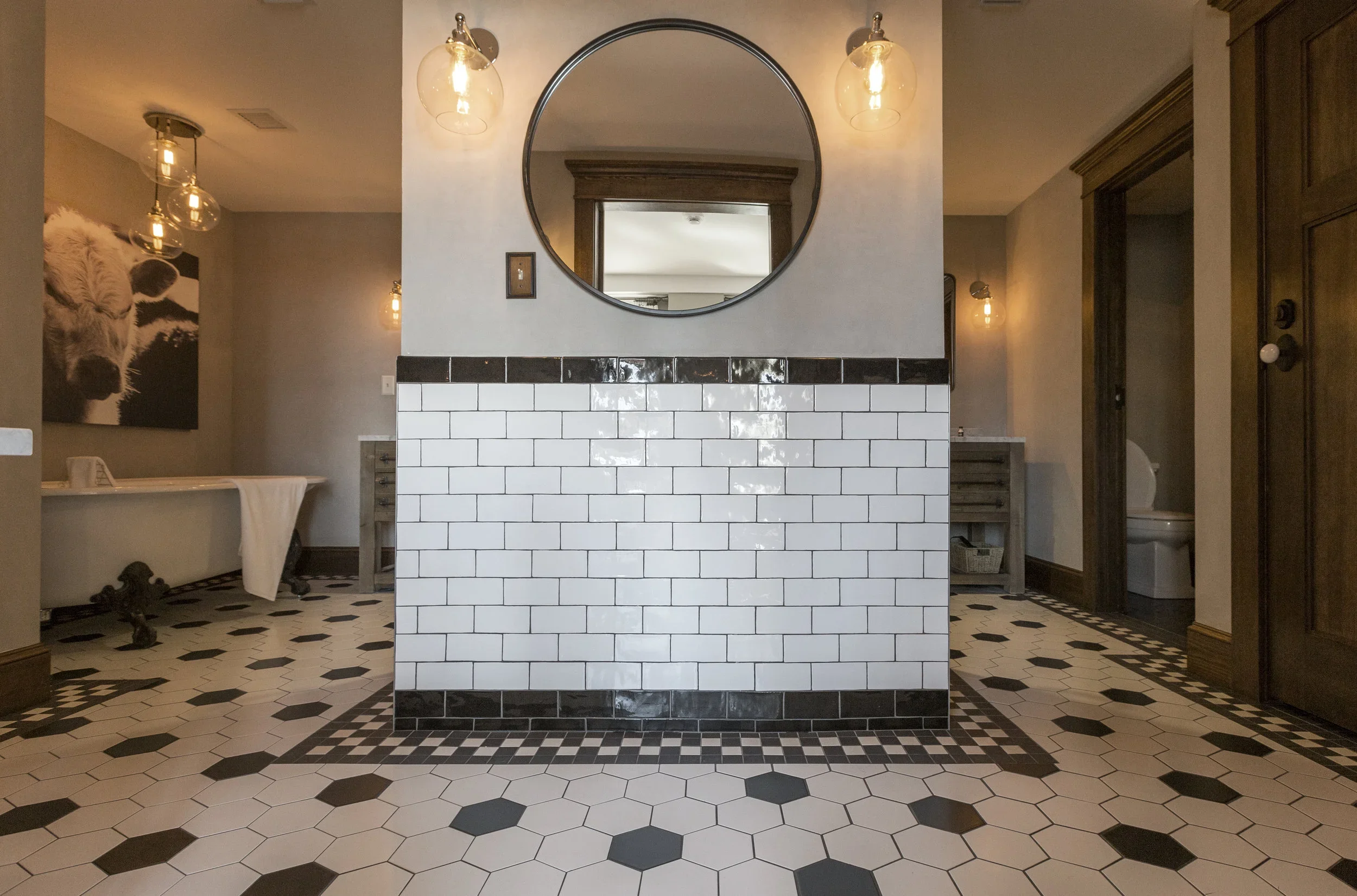 Interior of a modern restroom with black and white tile flooring, a half wall with white subway tiles and black trim, a round mirror above the wall, and vintage-style light fixtures. There is a bathtub on the left, and a toilet is visible on the righ