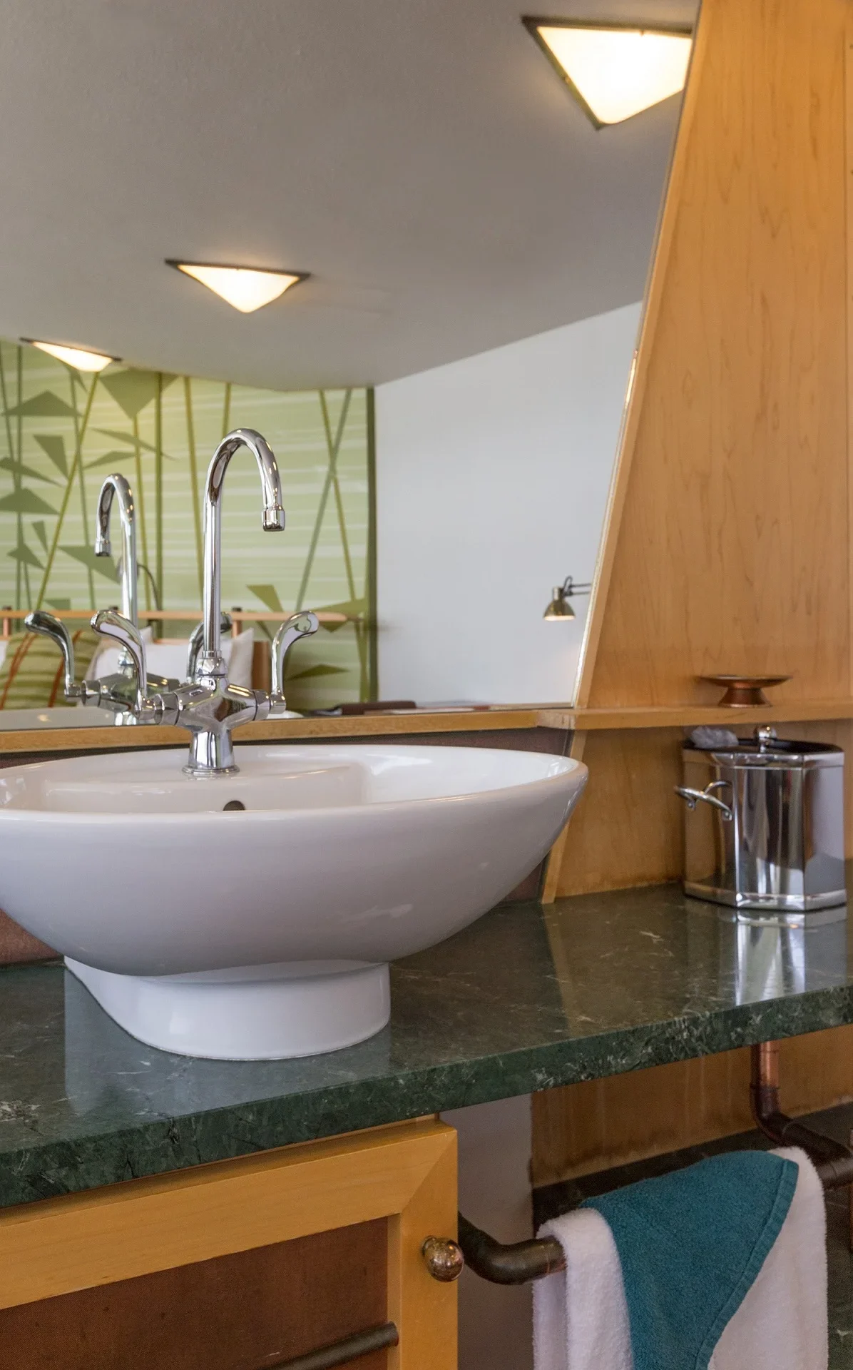 A bathroom sink area with a white vessel sink, chrome faucet, green marble countertop, wooden cabinetry, a mirror, a soap dispenser, and a towel hanging below.