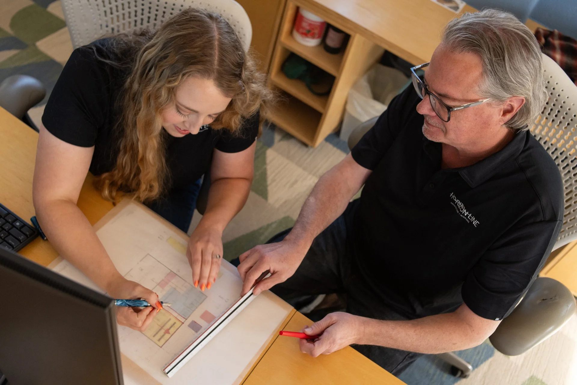 Two people, a woman and a man, are sitting at a desk looking at a color-coded architectural drawing or floor plan. They are engaged in a discussion, with the woman pointing at the plan and the man holding a red pen.