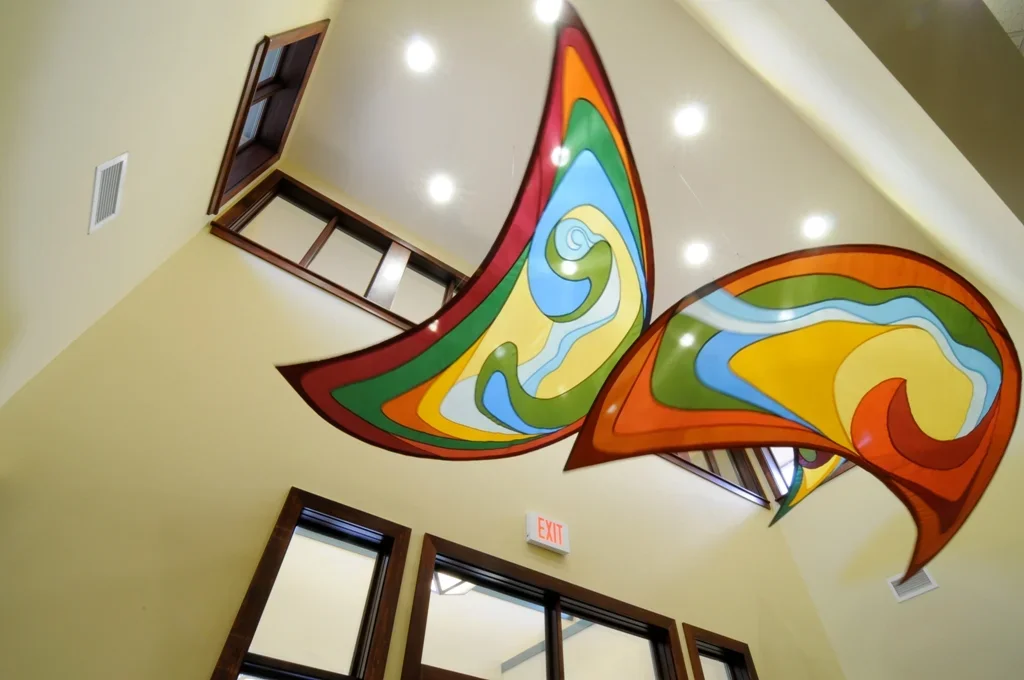 Colorful abstract stained glass decoration resembling butterfly or bird wings hanging from a high ceiling in an interior space.