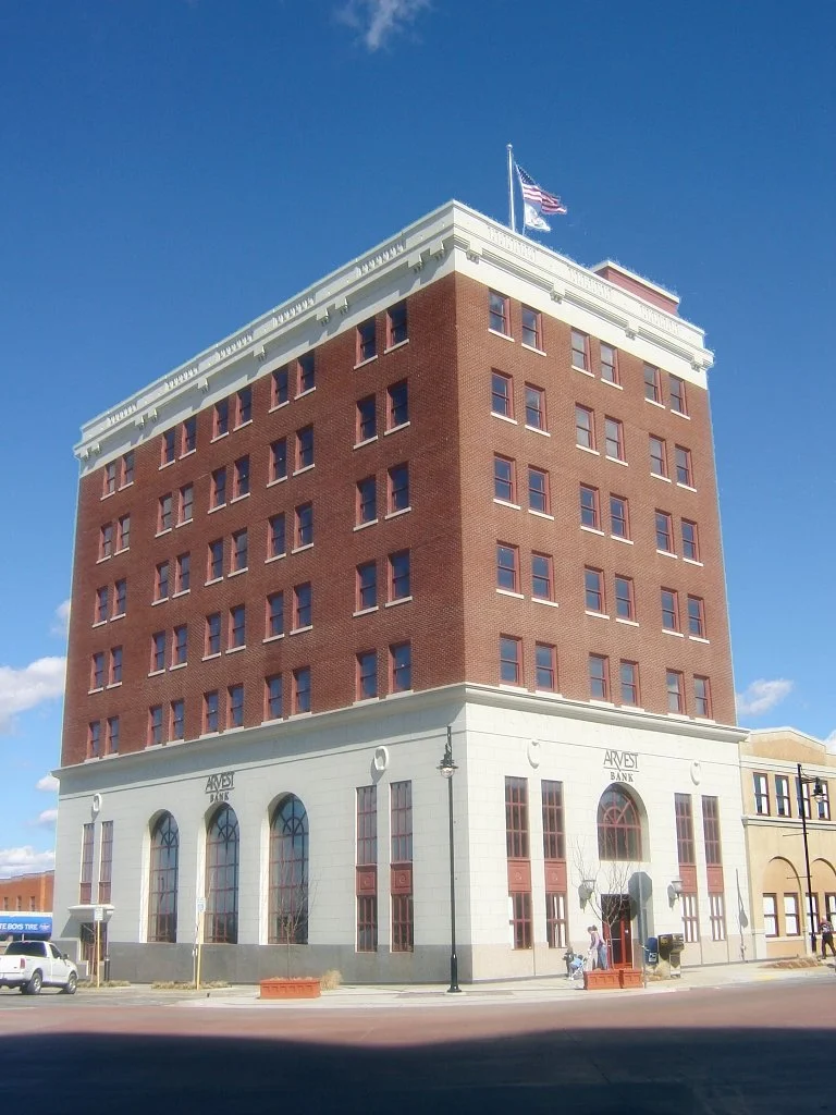 A tall brick building with a white base, labeled 'Arvest Bank,' with an American flag on the roof under a blue sky.