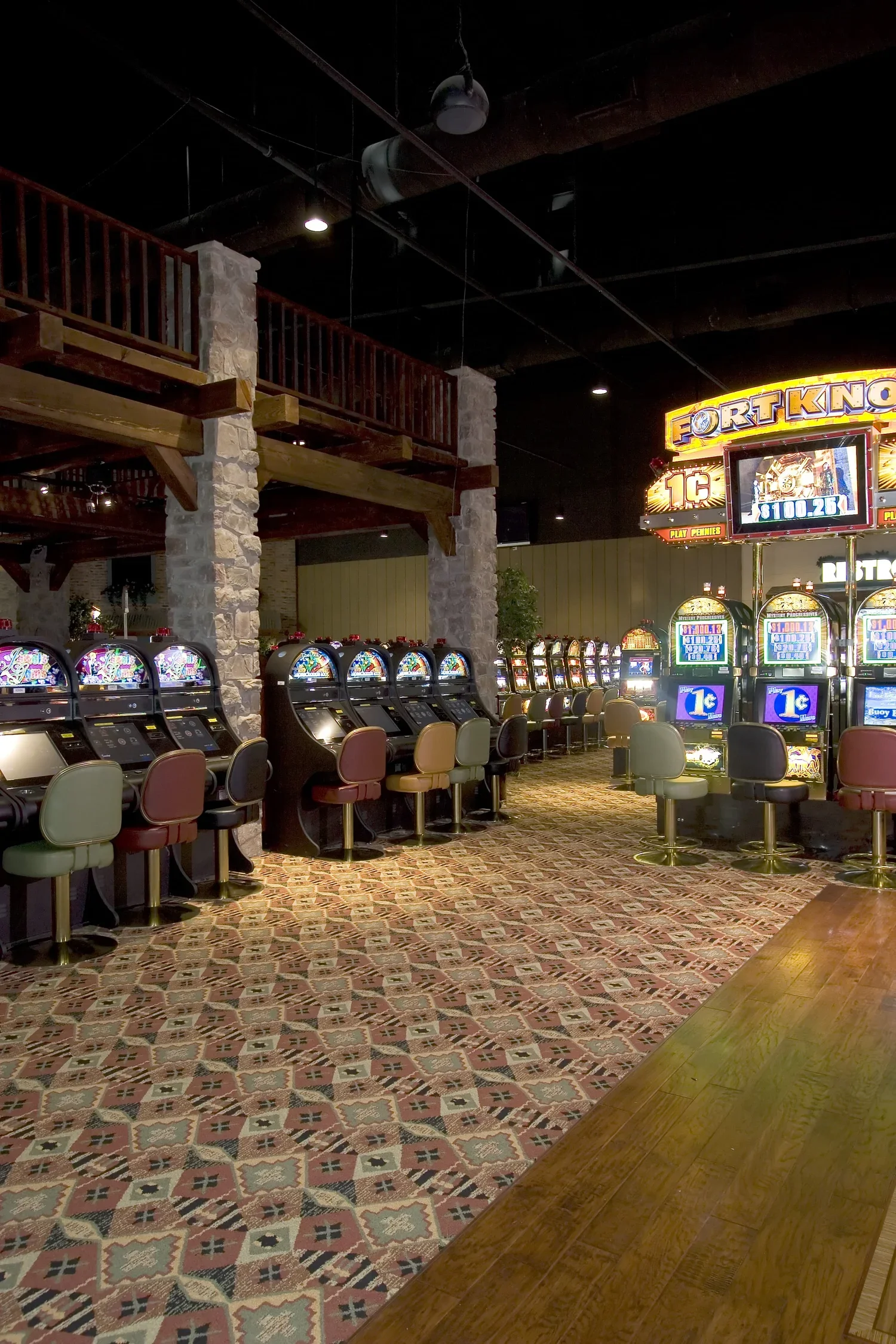 Interior of a casino with row of slot machines and chairs, stone pillars, wooden balcony, patterned carpet, and illuminated sign for fortunes.