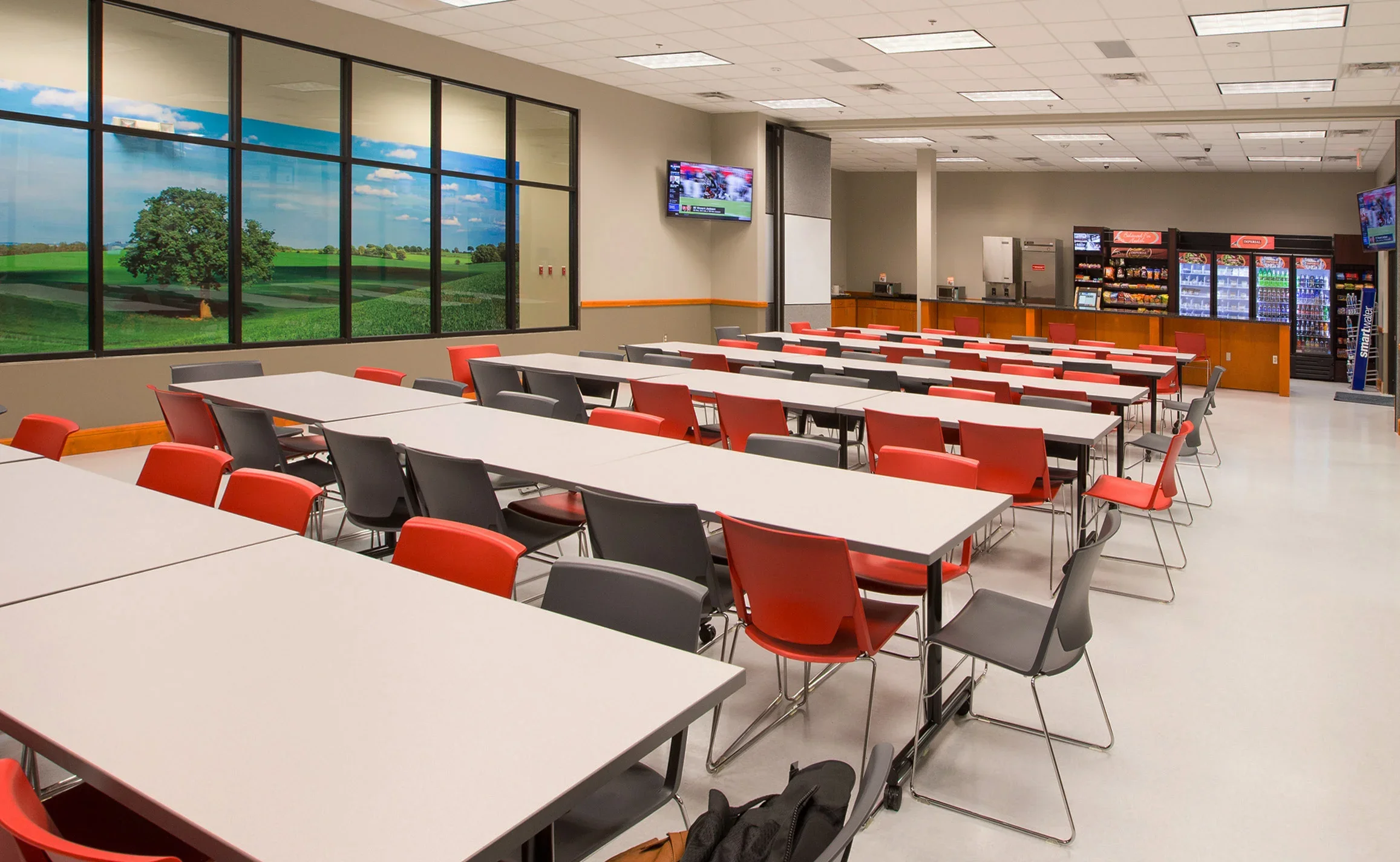 Empty classroom or conference room with white tables and red and gray chairs, large windows showing a green landscape with a tree and blue sky, mounted TV screens, and vending machines in the background.