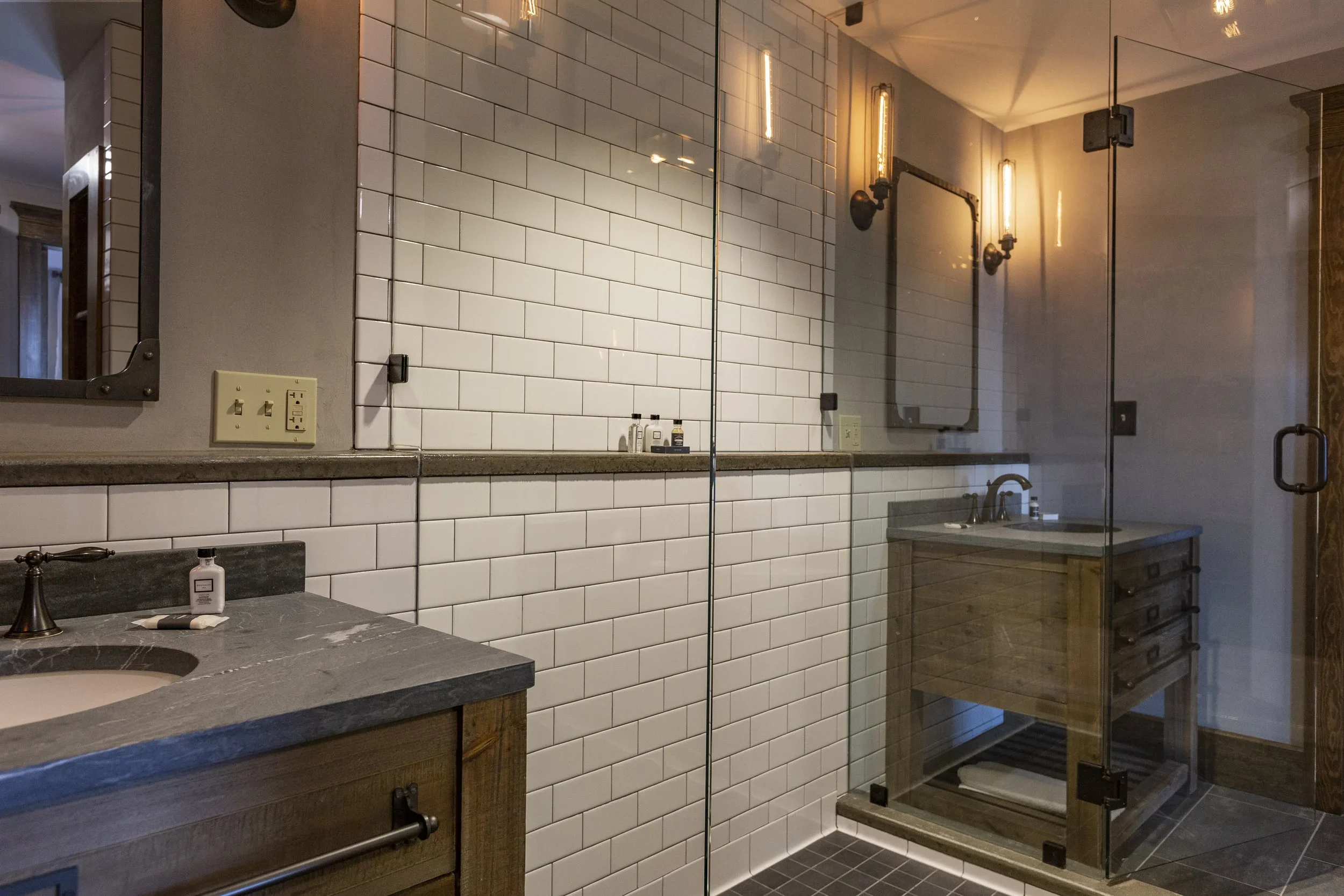 A bathroom with a wooden vanity, a marble countertop, a mirror, and a glass-enclosed shower with black hardware and white subway tiles.