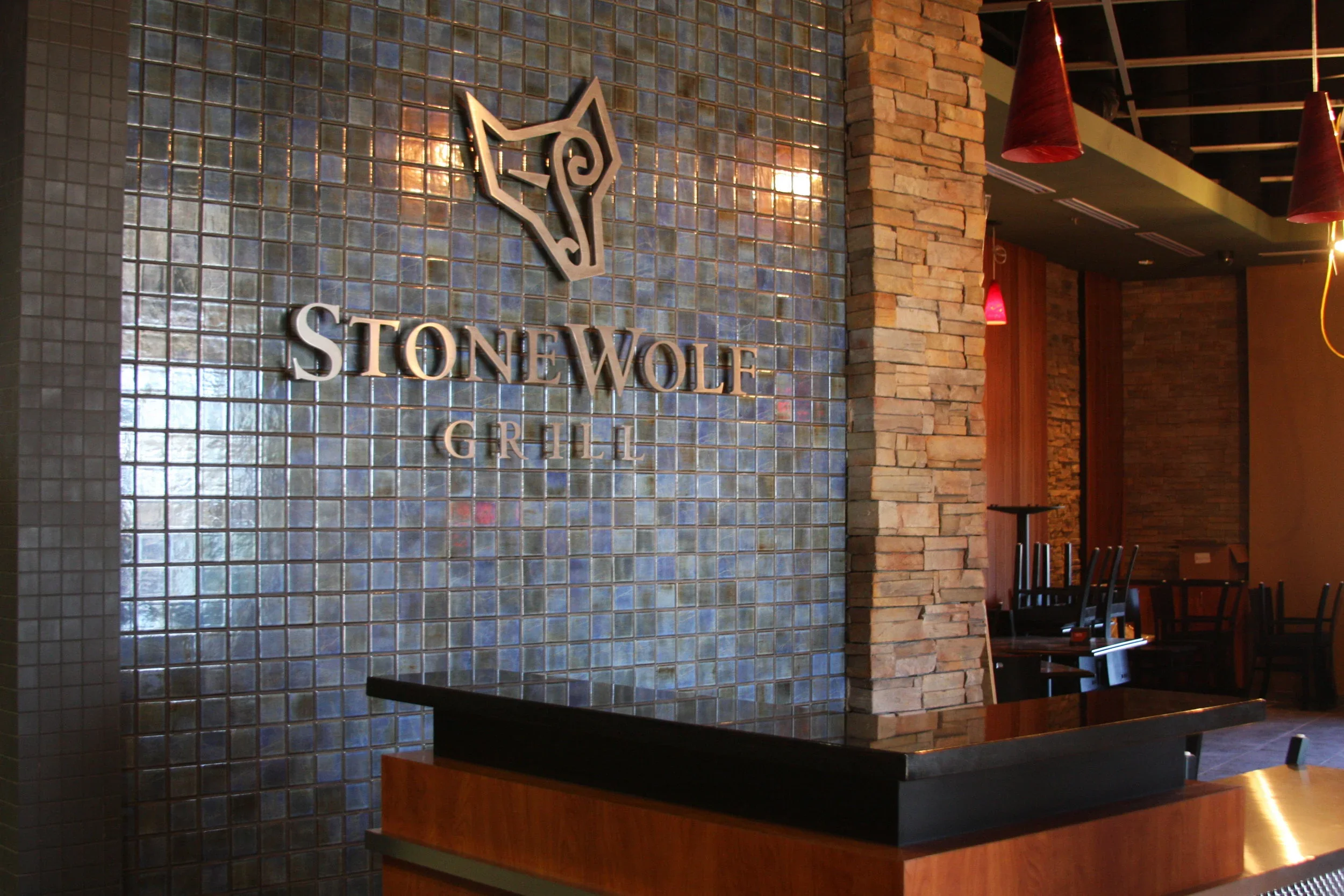 Interior of Stone Wolf Grill restaurant with a tiled wall featuring the logo and name, stone pillar, tables, chairs, and pendant lights.