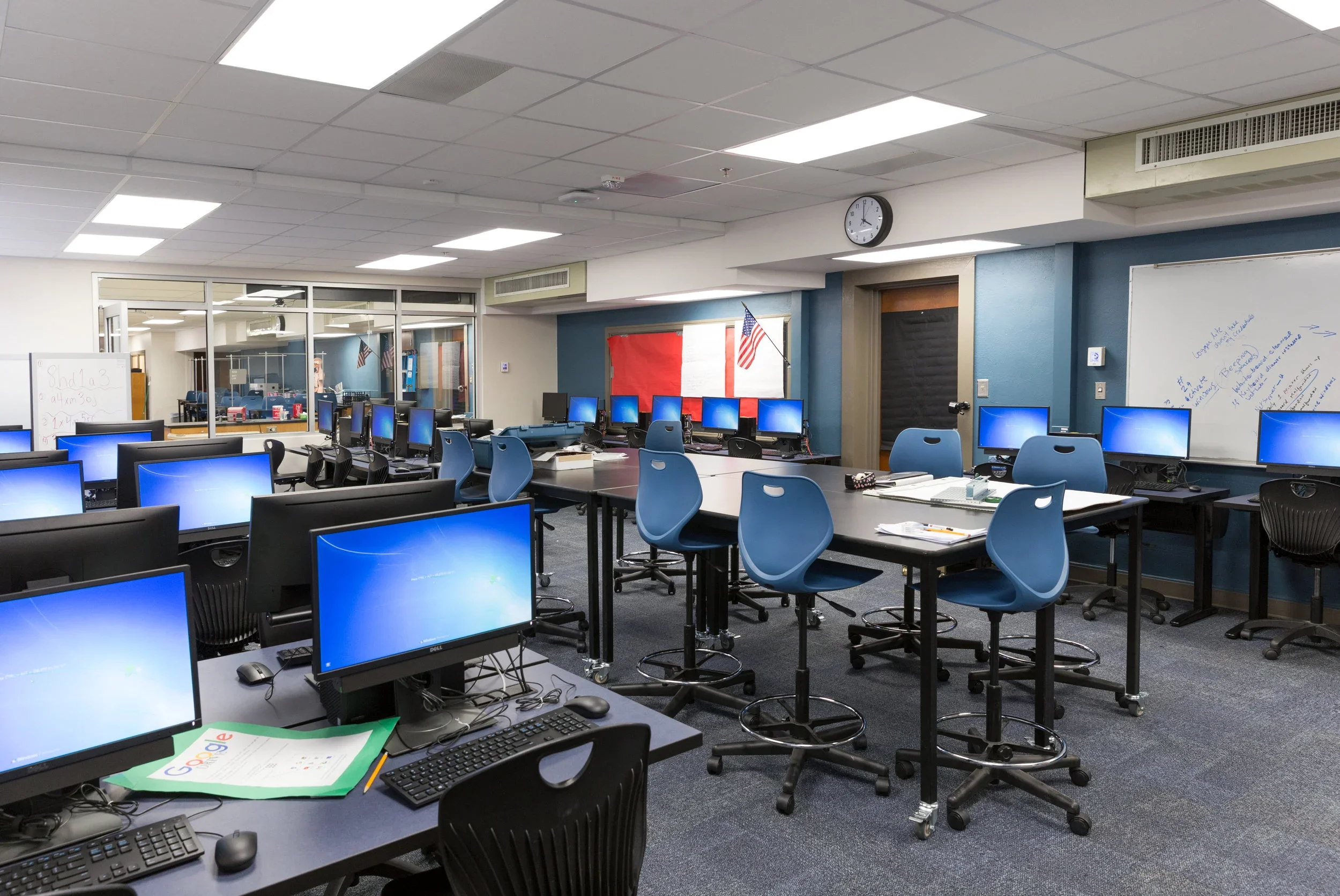 Empty computer lab classroom with multiple desktop computers on tables, blue chairs, whiteboard on the wall, and American flags.