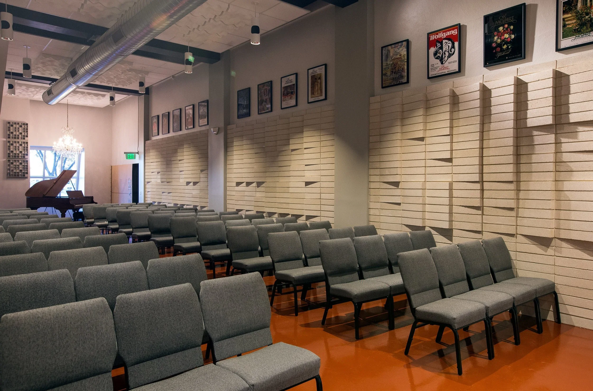 Empty concert hall with rows of gray chairs, a grand piano, framed posters on the walls, and a chandelier near the window.