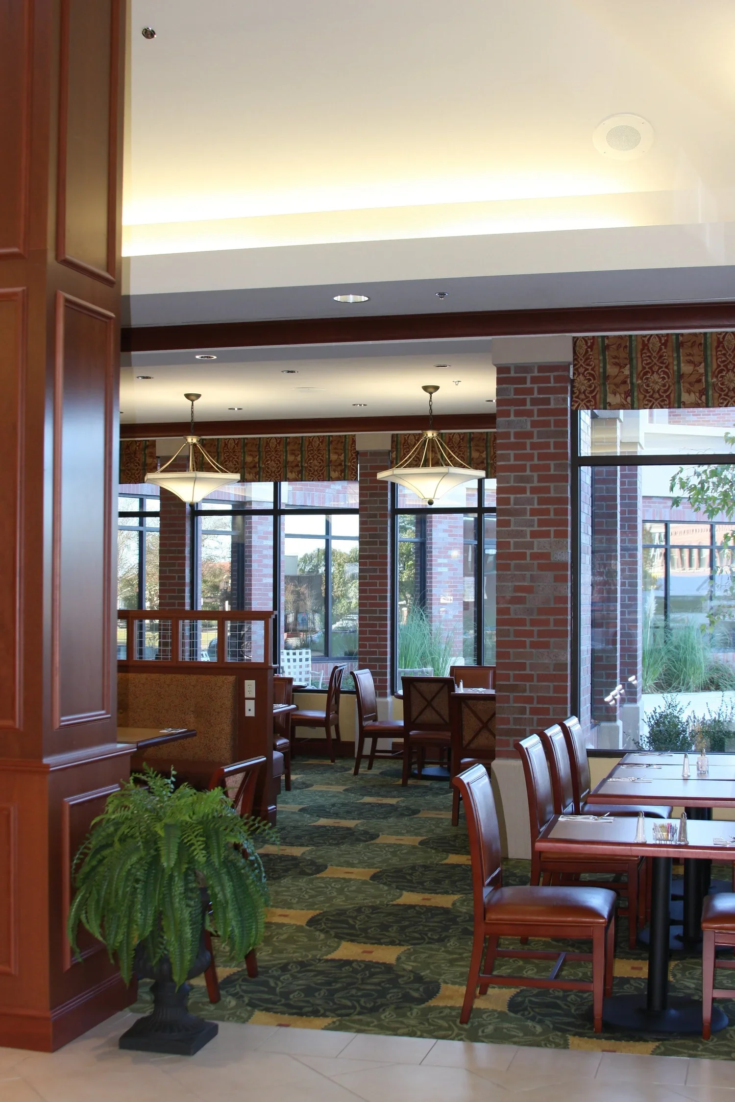 Interior view of a restaurant with red brick walls, large windows, wooden tables and chairs, hanging light fixtures, a green carpeting with floral patterns, and a potted fern plant in the foreground.