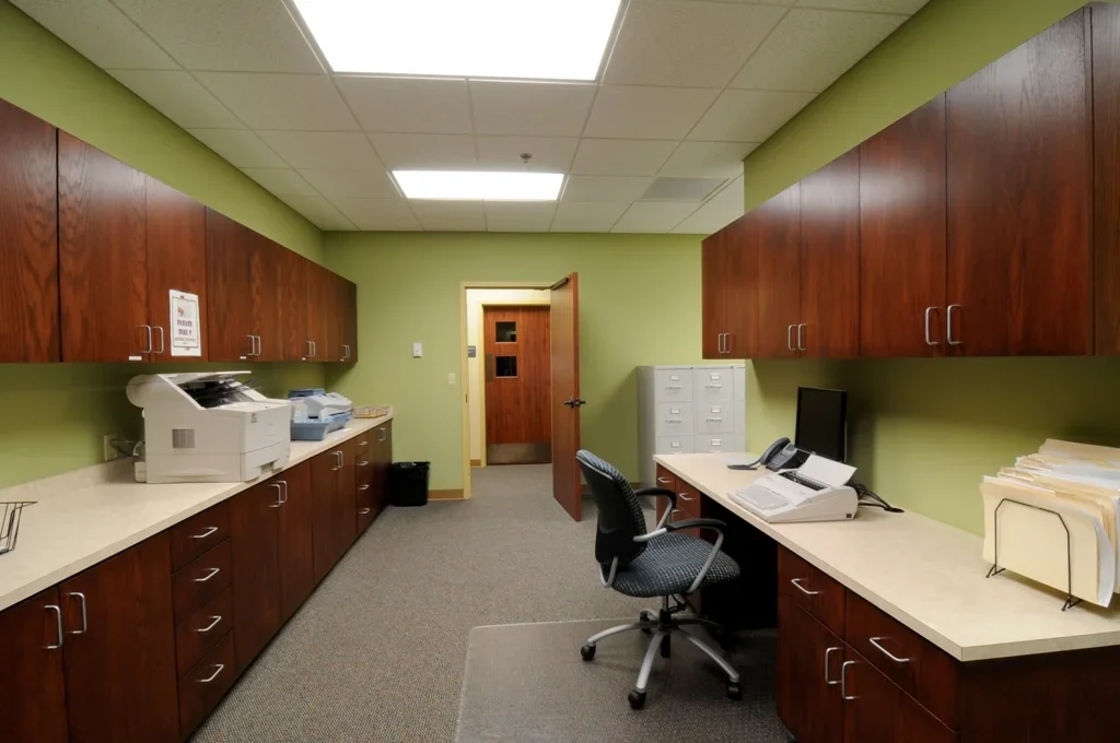 Office kitchenette or break room with wooden cabinets, a desk with a printer, phone, and papers, a filing cabinet, and various office supplies.