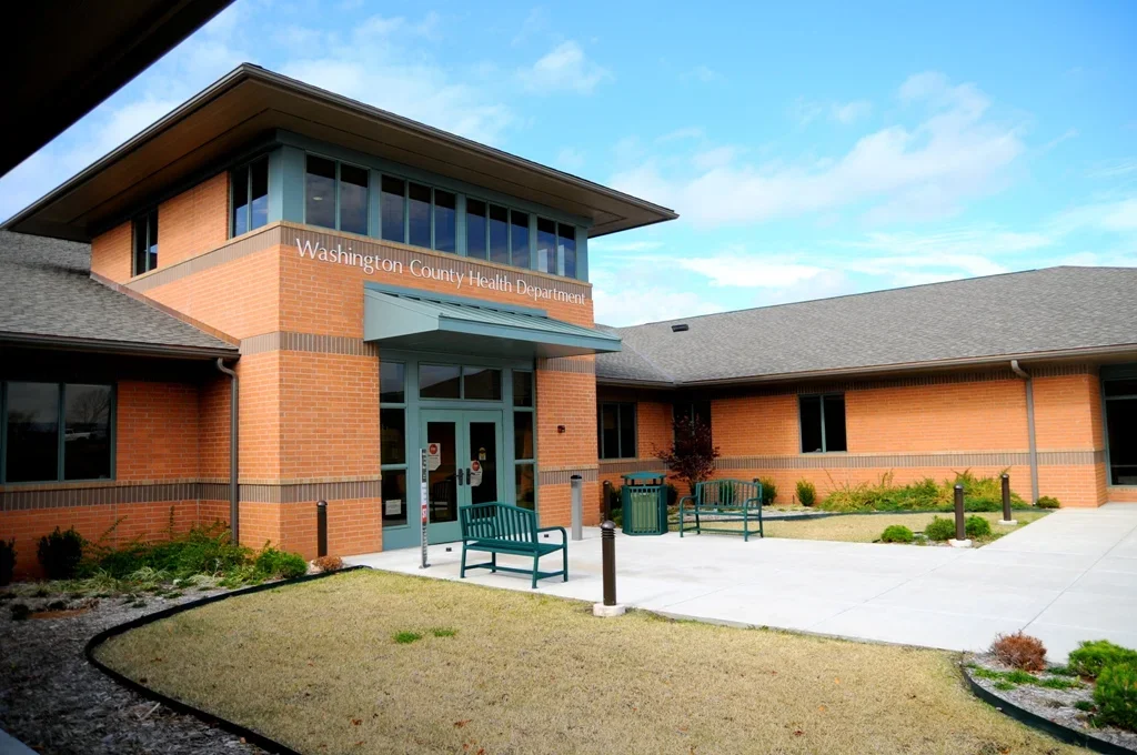 Washington County Health Department building with a brick exterior, glass windows, benches, and a landscaped entrance under a partly cloudy sky.