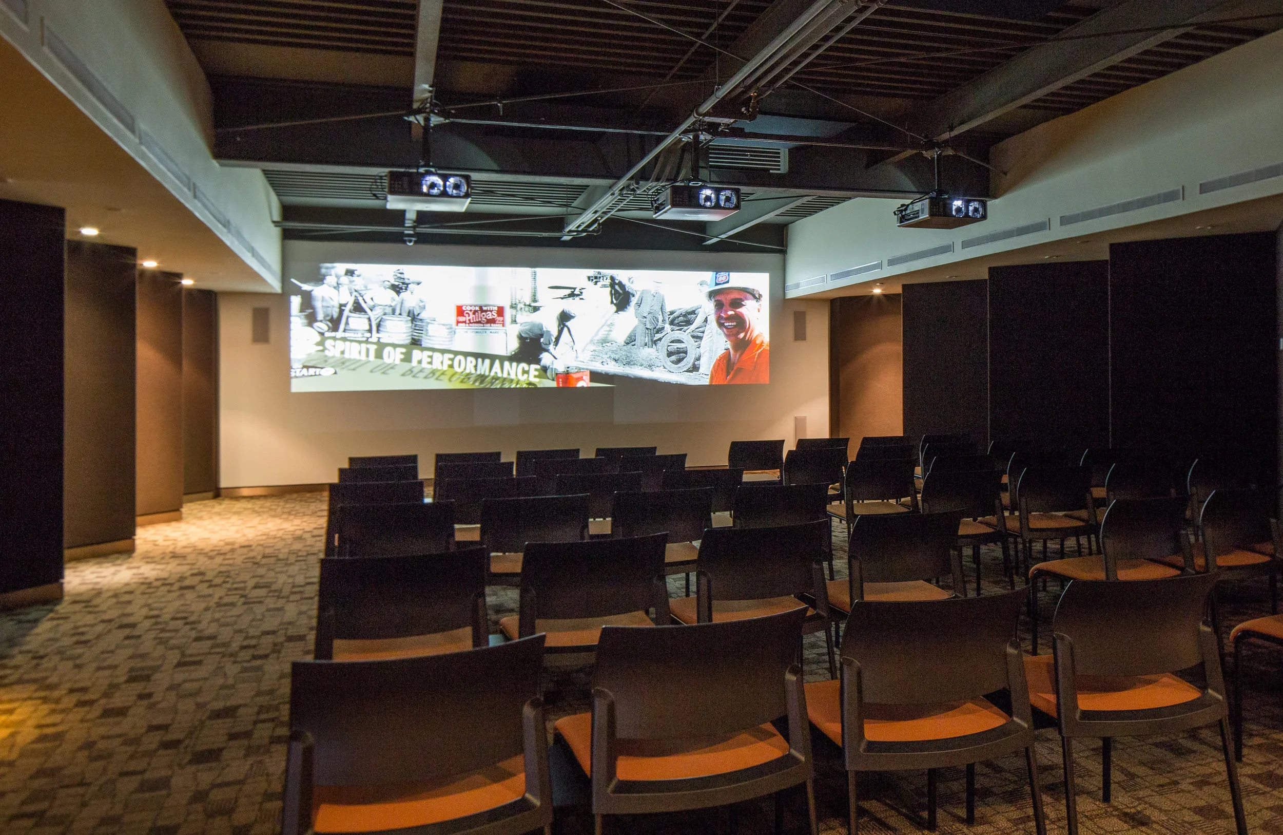 An empty movie theater with rows of black chairs facing a large screen displaying a black and white photo of workers and a smiling man in an orange shirt. The ceiling has black acoustic panels and mounted projectors.