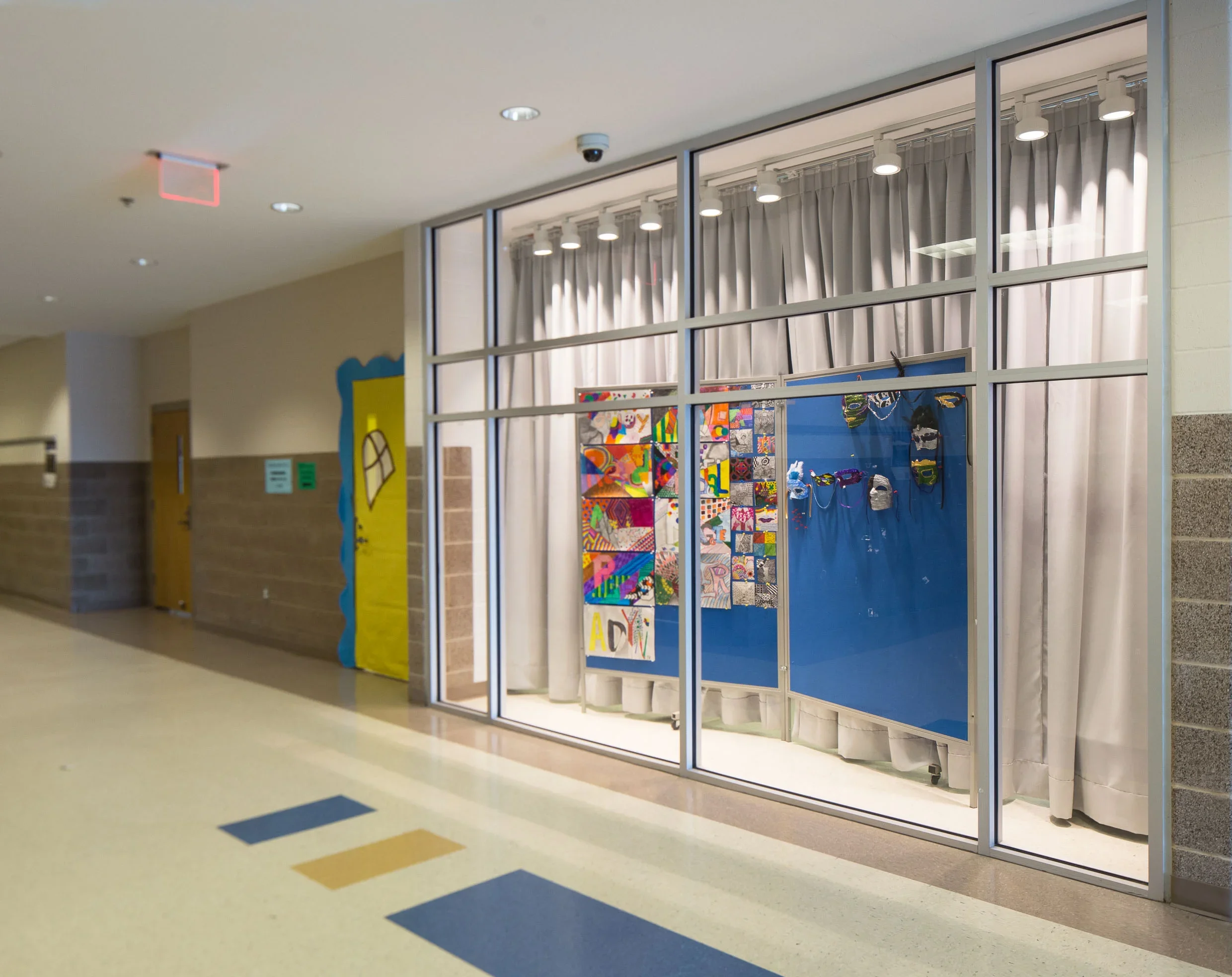 School hallway with a classroom display of student artwork behind glass windows, and a colorful door decoration on the wall.