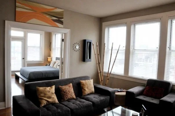 Living room with a dark gray sofa decorated with multiple pillows, next to a large window with blinds. Behind the sofa, there's a smaller armchair with pillows, a side table, and a stack of wooden sticks or blinds leaning against the wall. In the bac