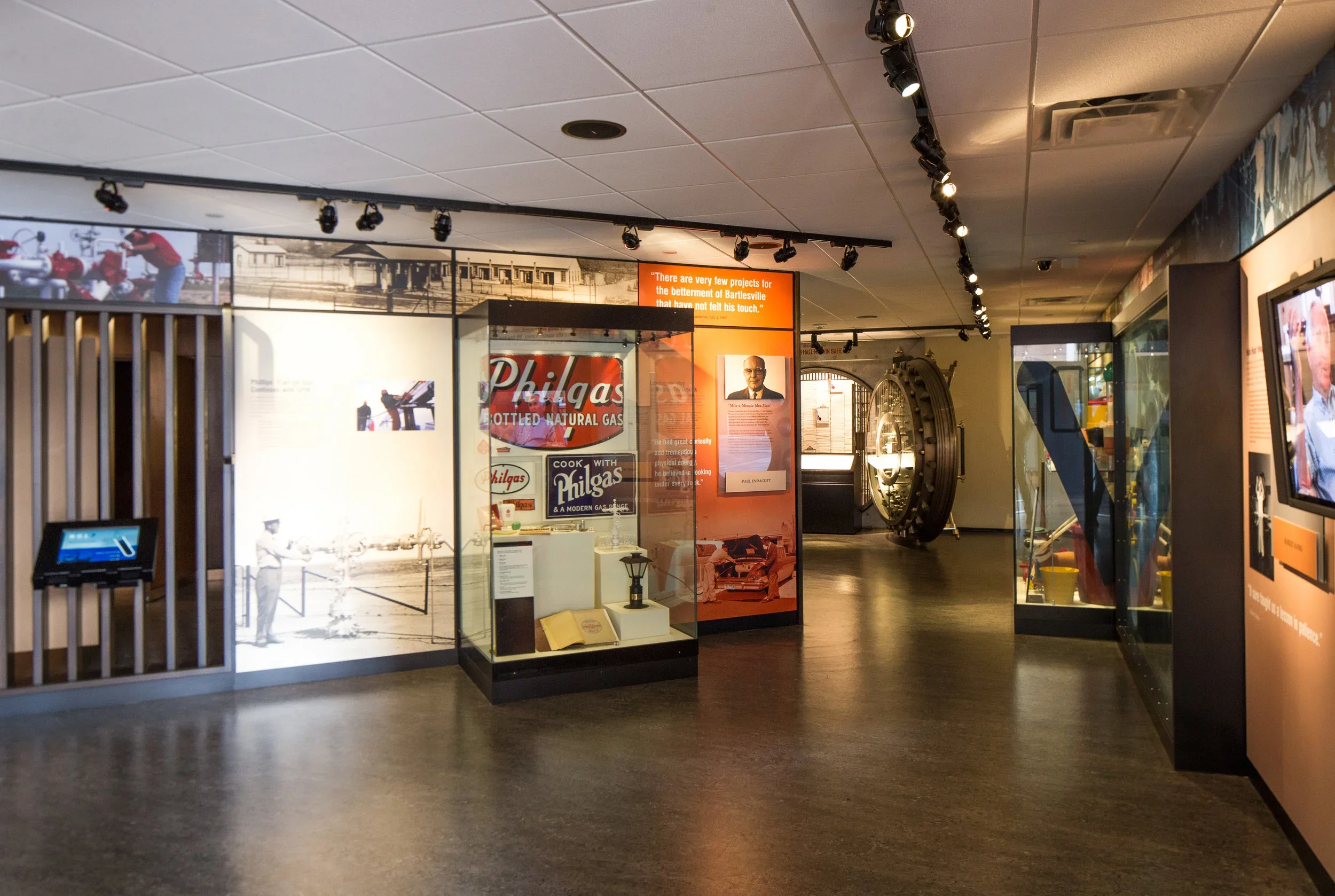 An interior view of a museum exhibit with display cases, informational panels, and a large vault door in the background. The floor is dark wood, and the ceiling has track lighting.