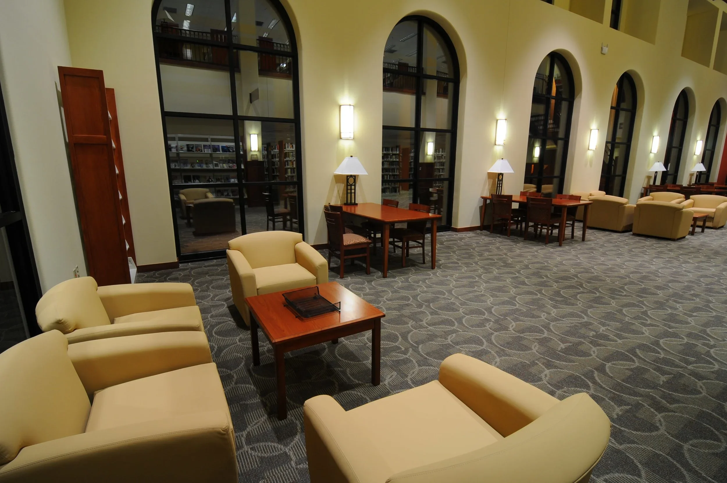 A spacious library lounge with beige armchairs, wooden coffee tables, and standing lamps, large arched windows, and bookshelves visible through glass doors.
