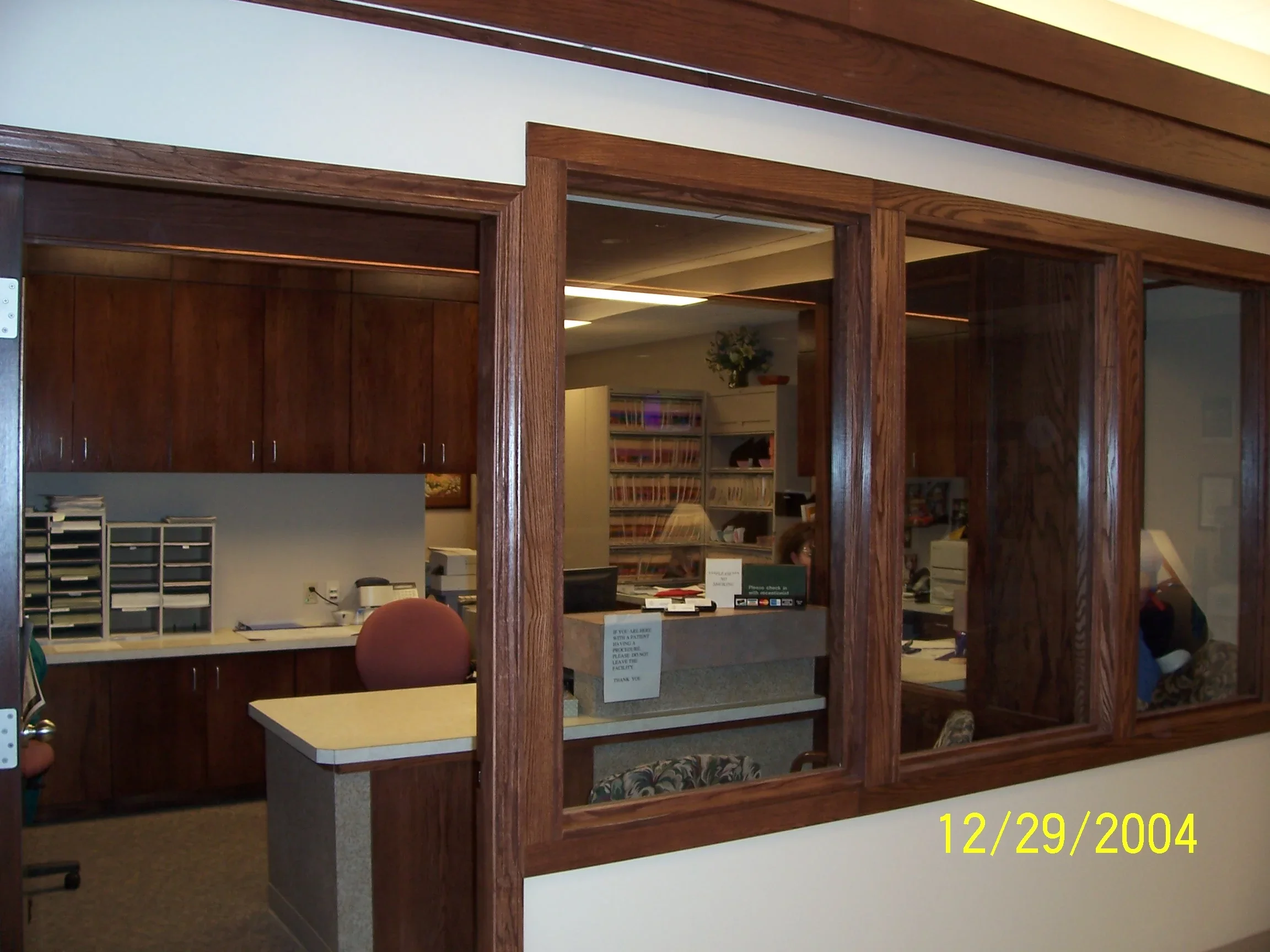 View of an office reception area seen through a large window with wooden framing, showing a desk with a computer, office chairs, shelves filled with files, and a person working at a second desk.