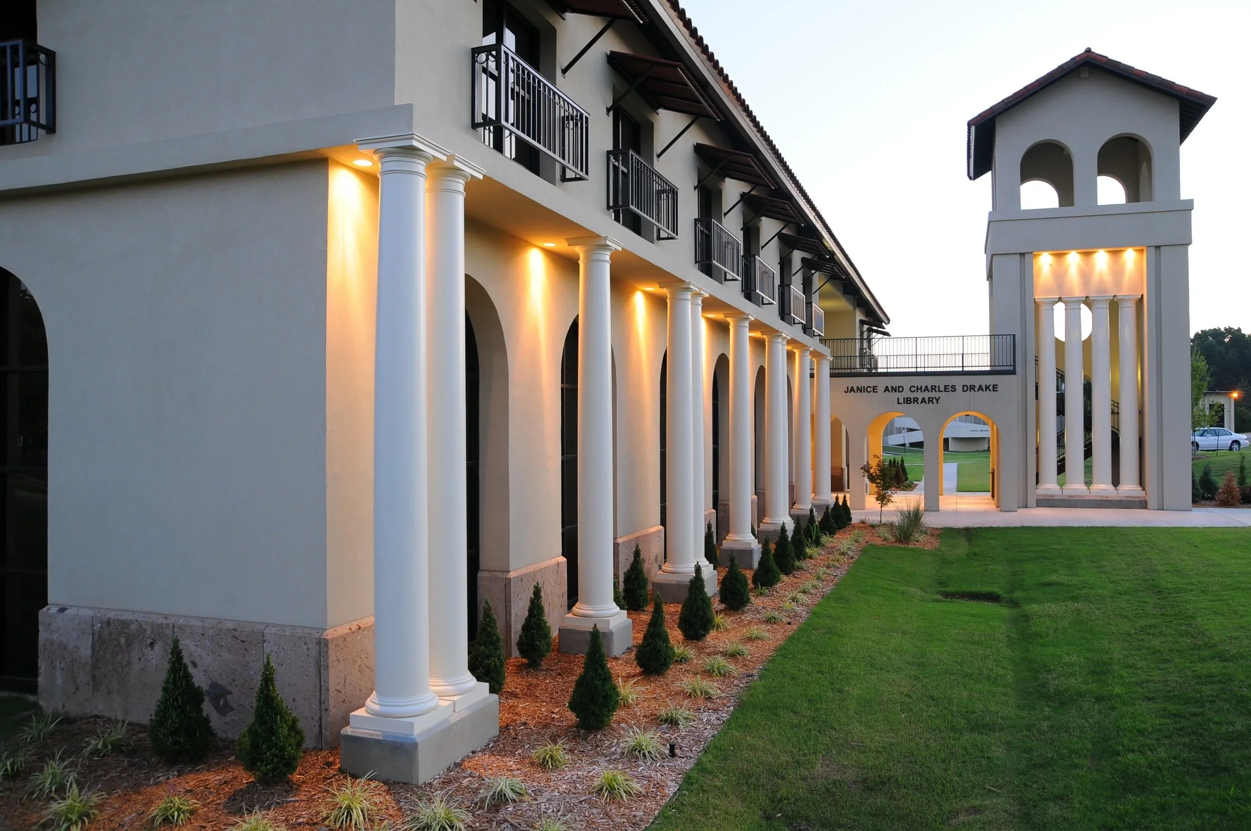 Exterior of the Janice and Charles Drake Library with lit columns, black balconies, a green lawn, and a bell tower at dusk.