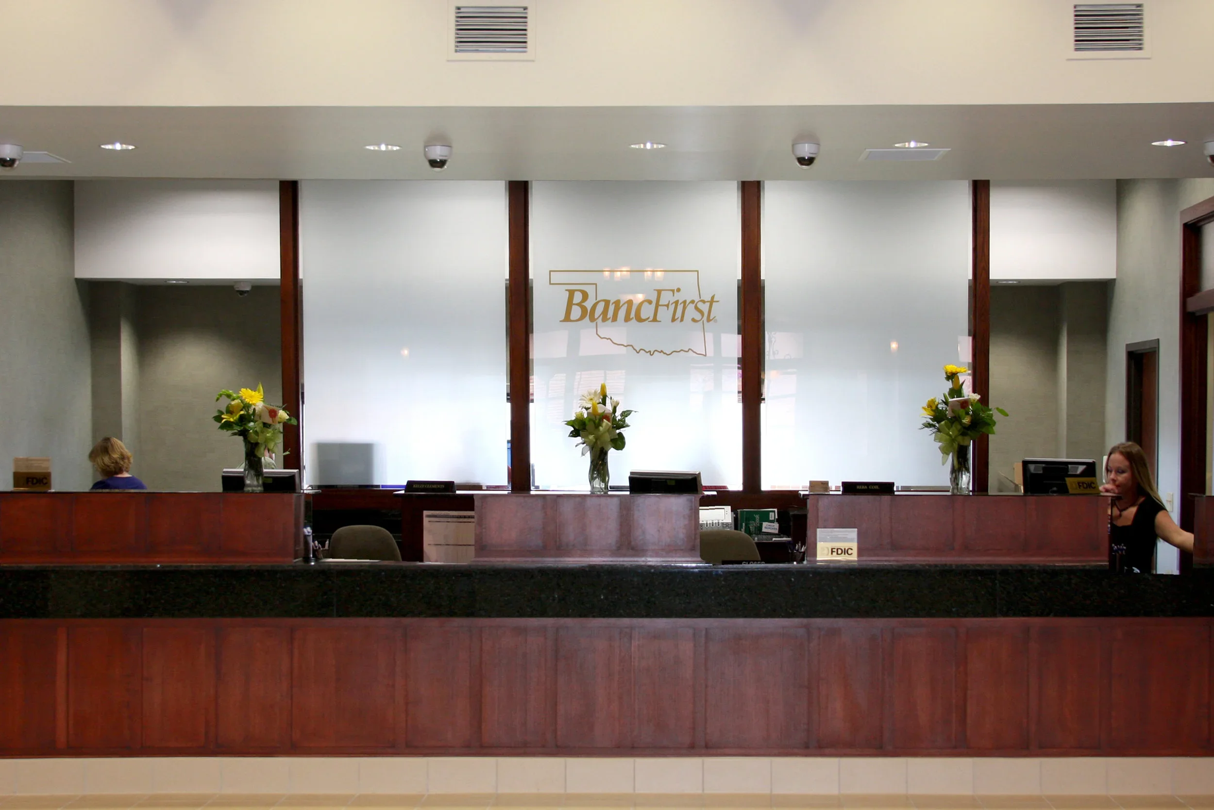 Lobby reception area with a wooden desk, three flower vases, and the logo 'BancFirst' on the frosted glass behind the desk.