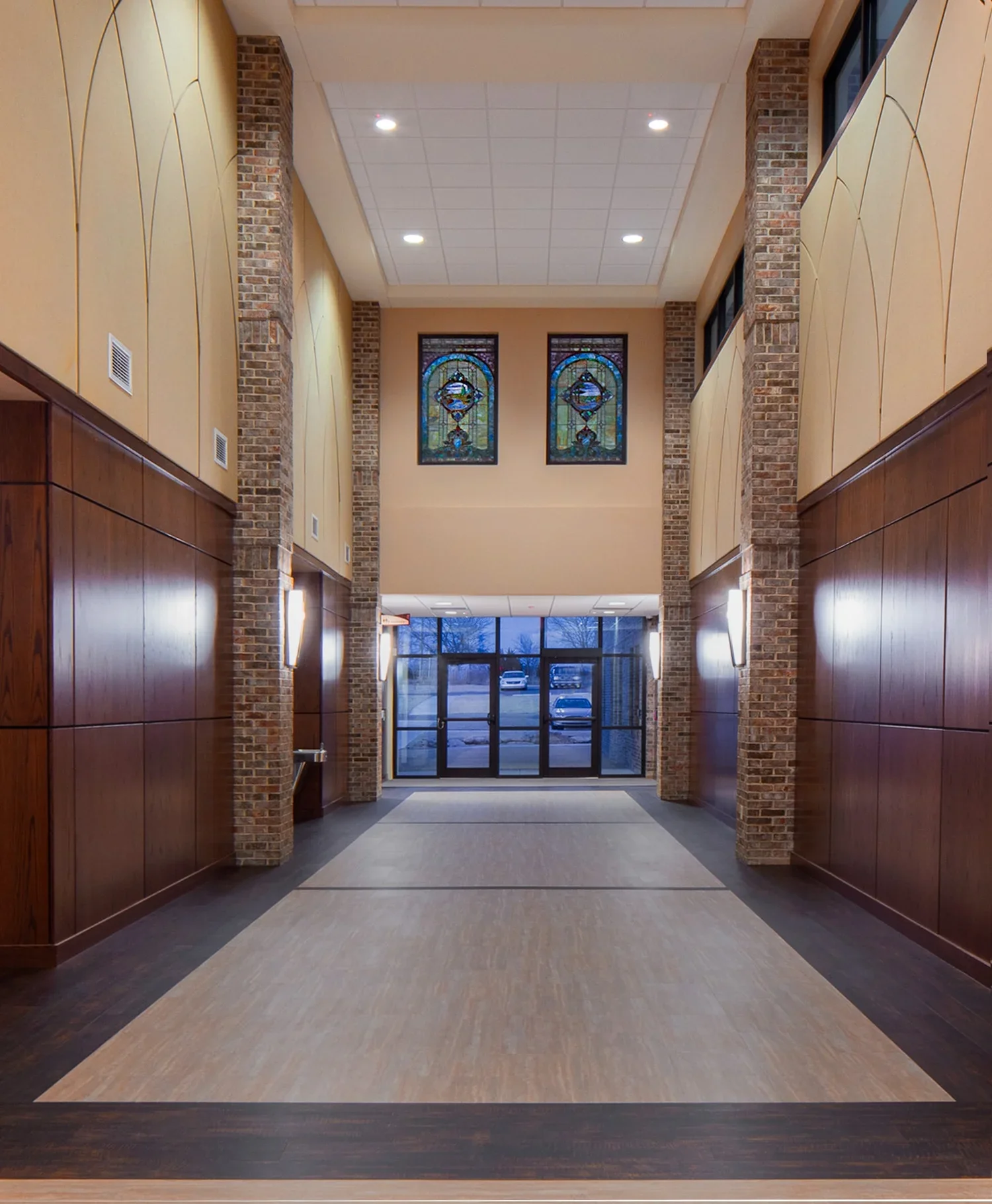 Interior view of building entrance with glass doors, brick columns, and stained glass windows, wood-paneled walls, and a tiled floor.