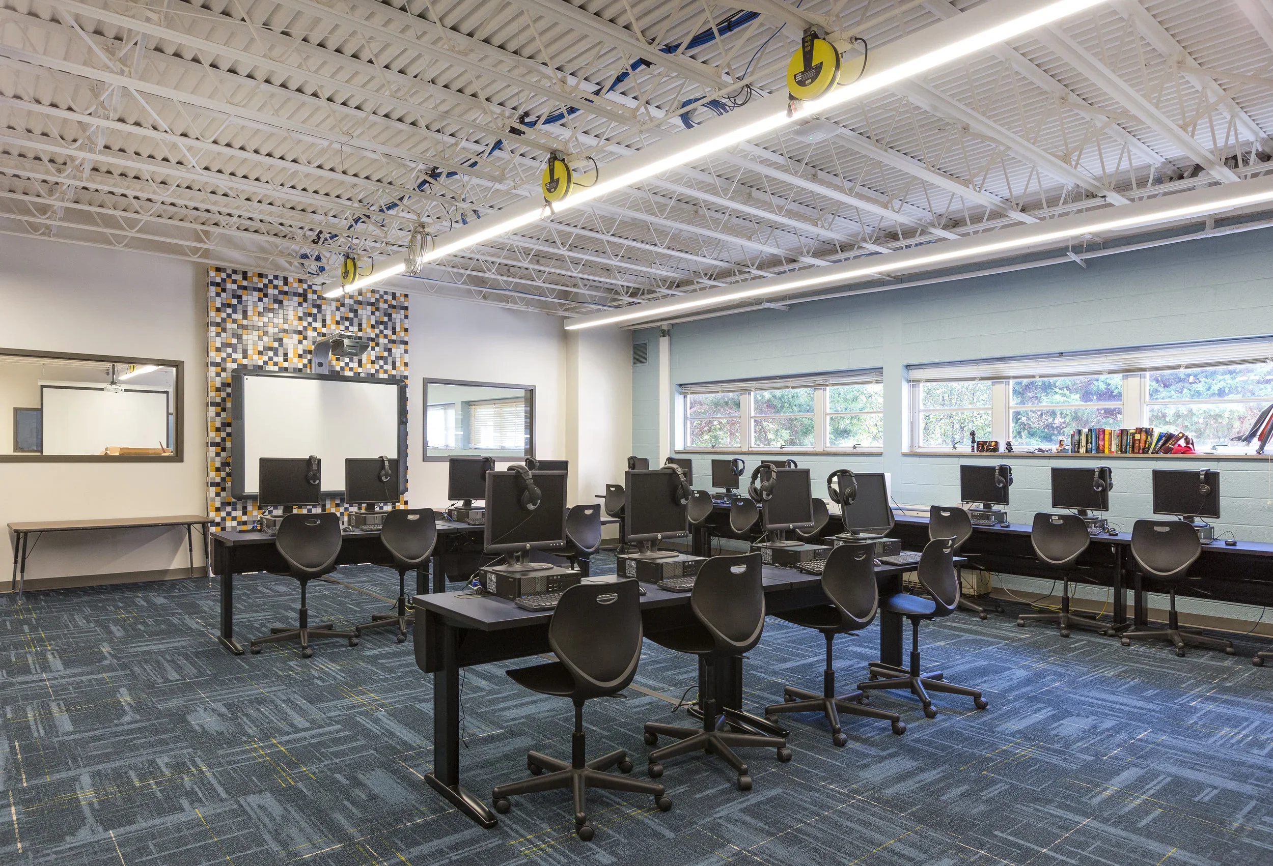 A classroom with multiple computer stations, each with a monitor, keyboard, and headphones, arranged on long black tables. There are black chairs and large windows on the right side of the room. The ceiling has exposed white beams and hanging yellow 
