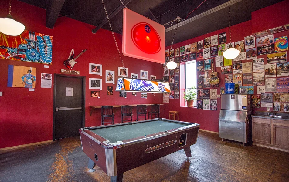 A red-walled room with a pool table in the center inside Buford Lofts, surrounded by vintage posters, photos, and artwork, with hanging lights and a neon sign, creating a cozy bar or game room atmosphere.