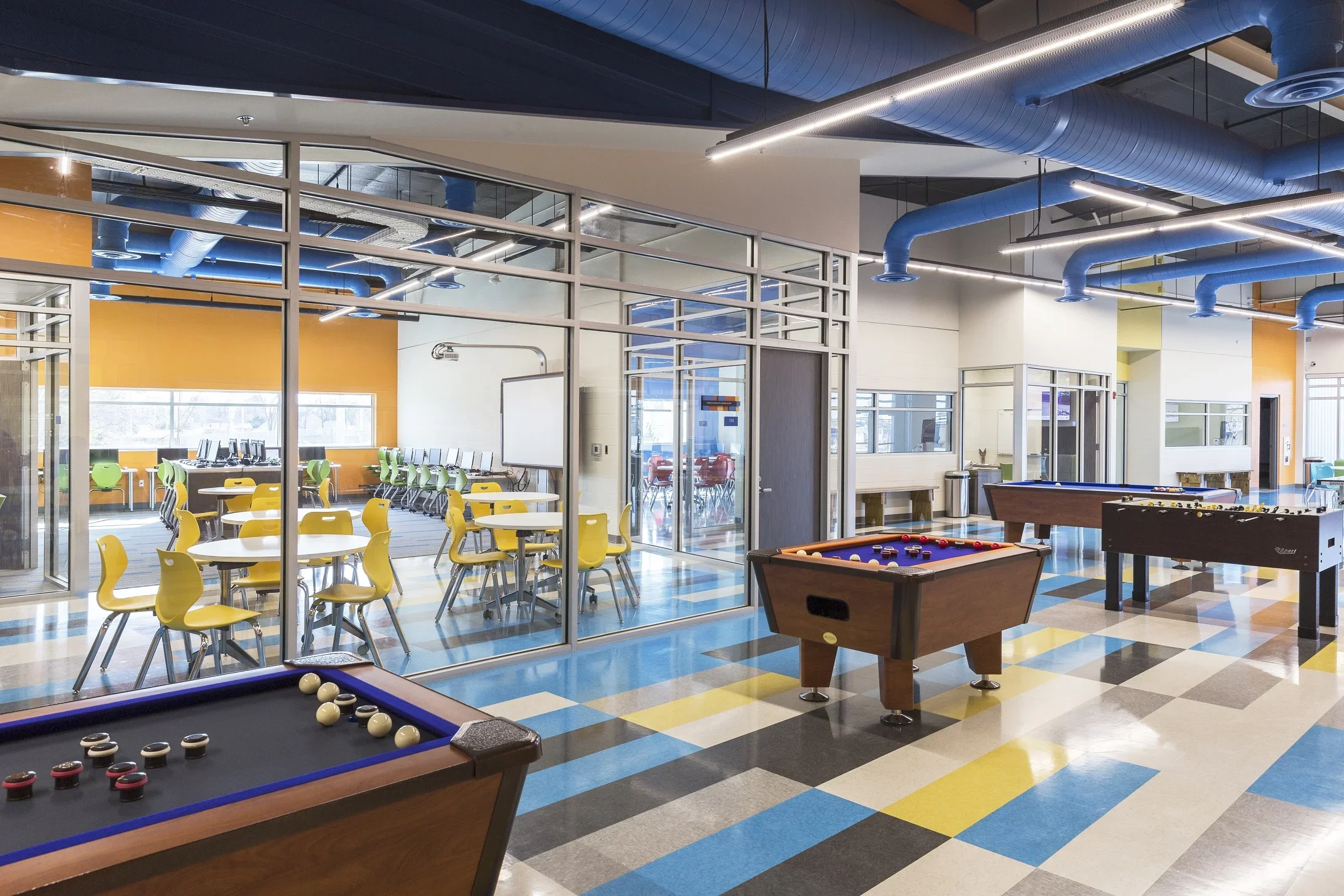Indoor recreational space with colorful tiled floor and multiple pool tables. Behind a glass wall, there is a classroom or computer lab with yellow chairs and tables, as well as a row of computers against the window. The ceiling has exposed blue duct