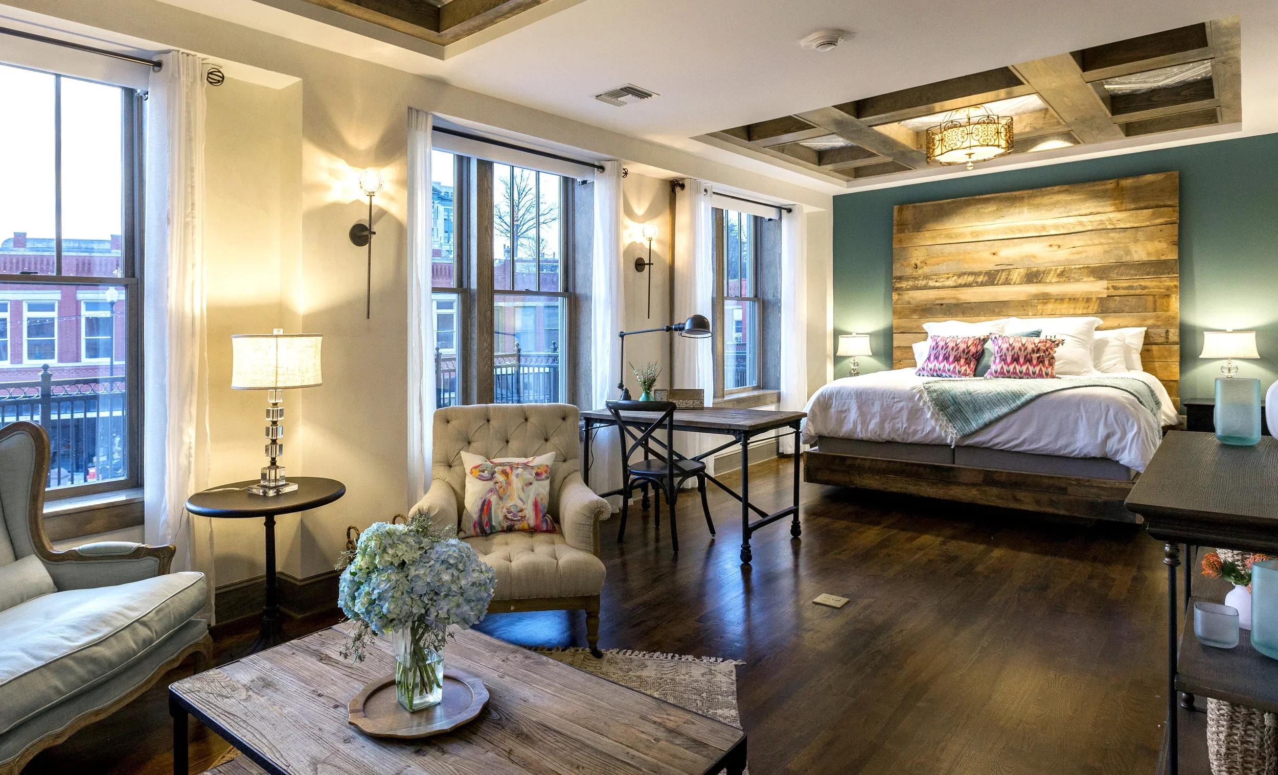 A modern bedroom with a rustic wooden headboard, white bedding, and colorful pillows. The room features three large windows with white curtains, a cozy armchair with a colorful pillow, a wooden coffee table with a flower arrangement, and a work desk 