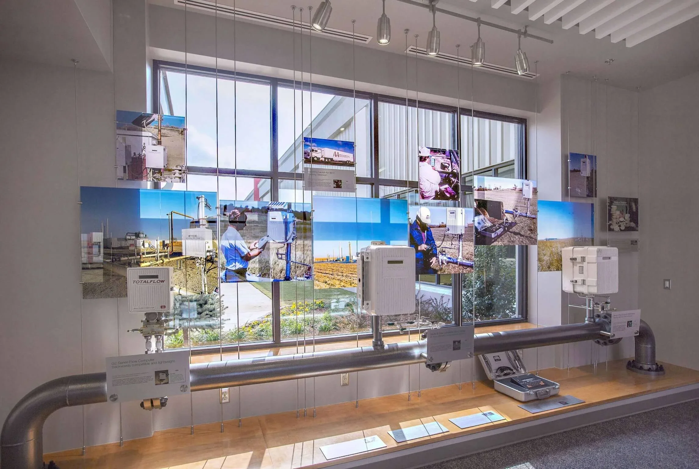 Exhibit displaying industrial equipment and photographs of fieldwork through a large window, with pipes and monitors in the foreground.