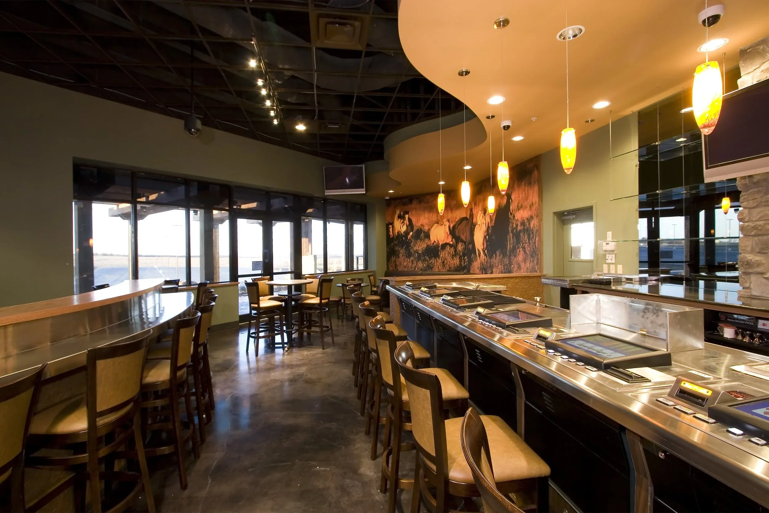 Interior of a bar or restaurant with bar stools, a long bar counter, hanging pendant lights, decorative horse wall art, and large windows letting in natural light.