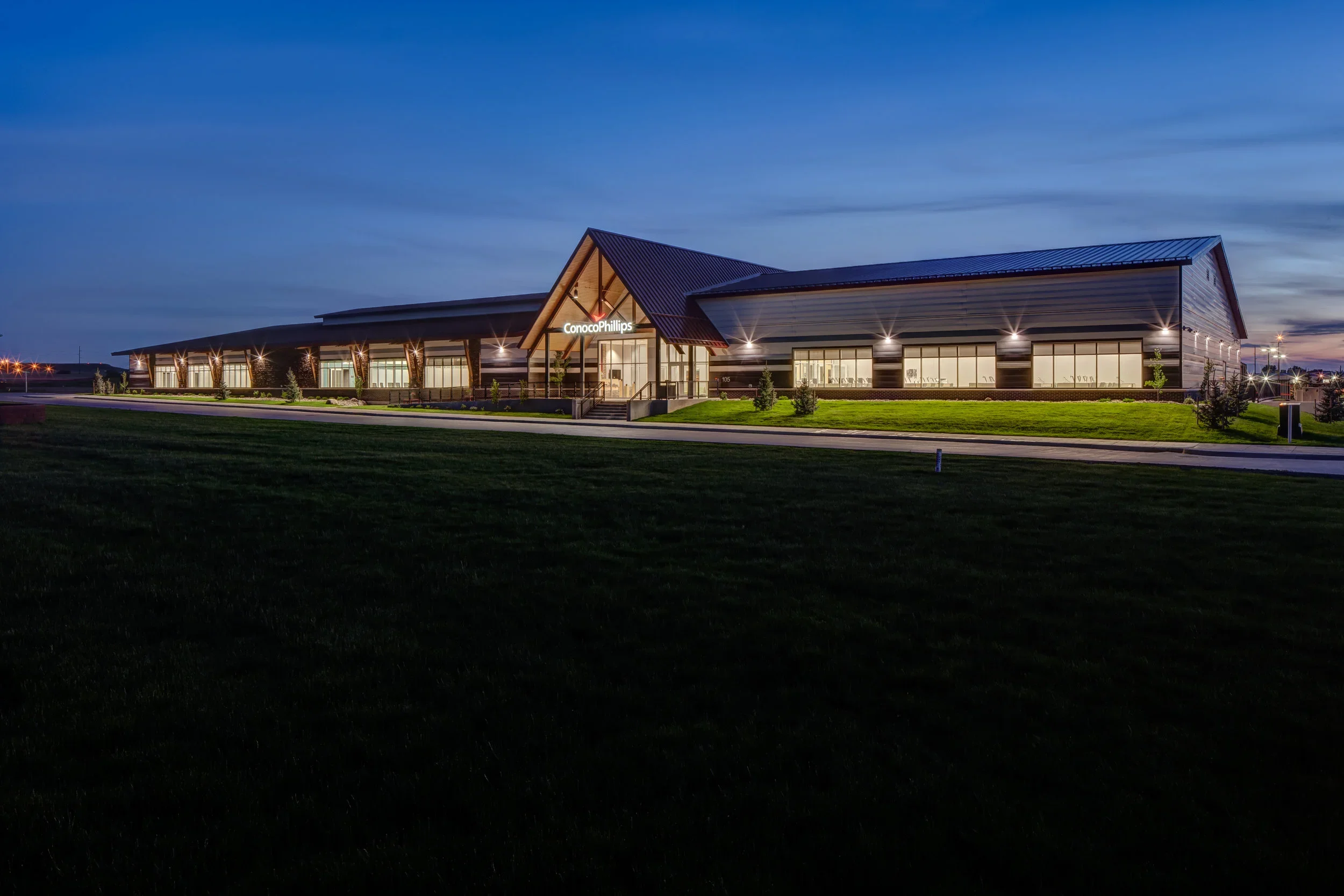 Exterior view of a modern office building with the sign 'ConocoPhillips', illuminated at dusk, surrounded by well-maintained grass and a sidewalk.