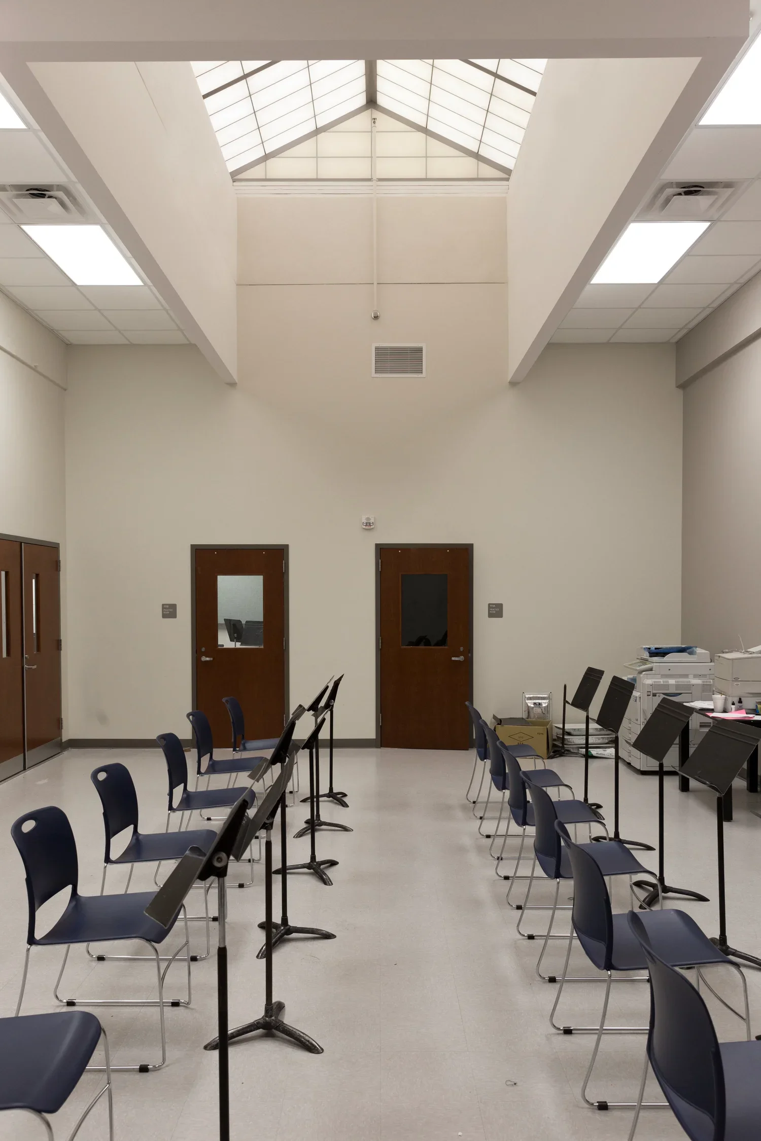 Empty music practice room with chairs and music stands, two doors with windows, and a high ceiling with skylight.