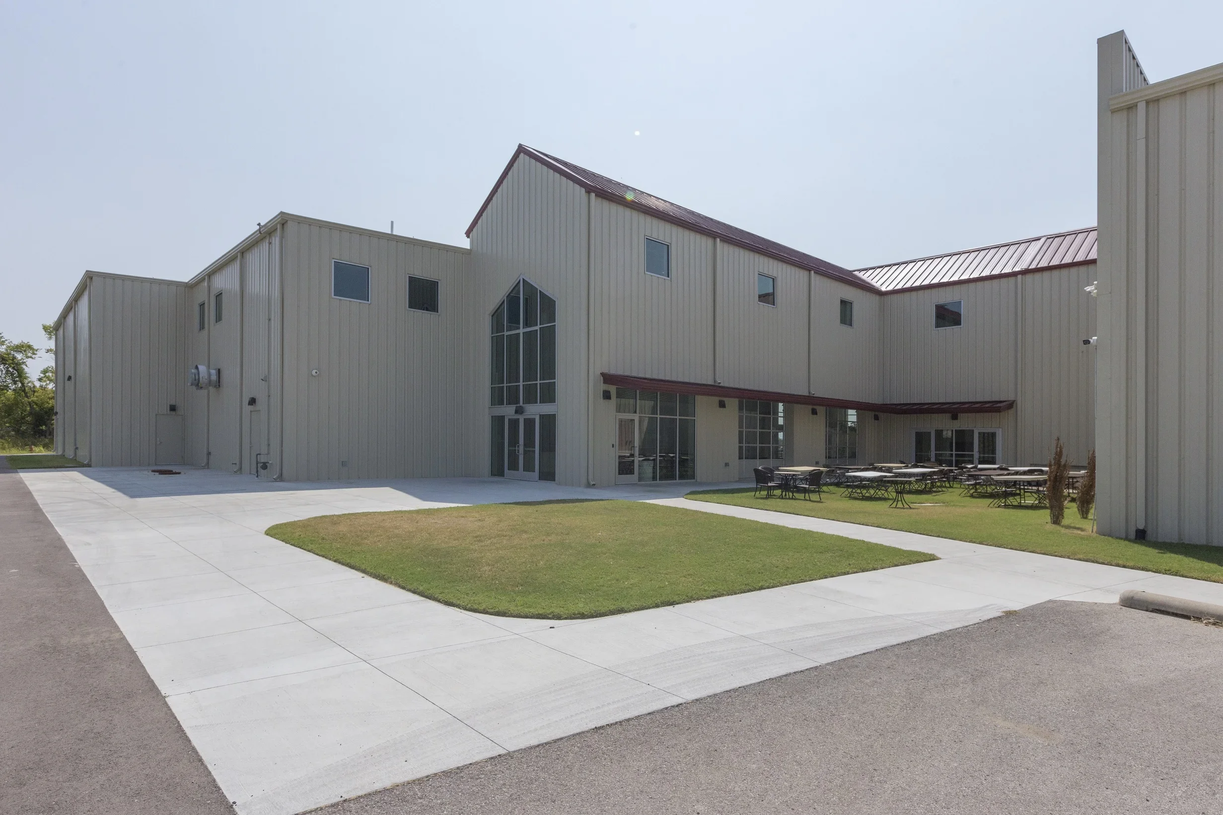 Exterior of a modern, beige metal building with multiple levels, large glass entrance, outdoor seating area, and landscaped lawn with walkways.