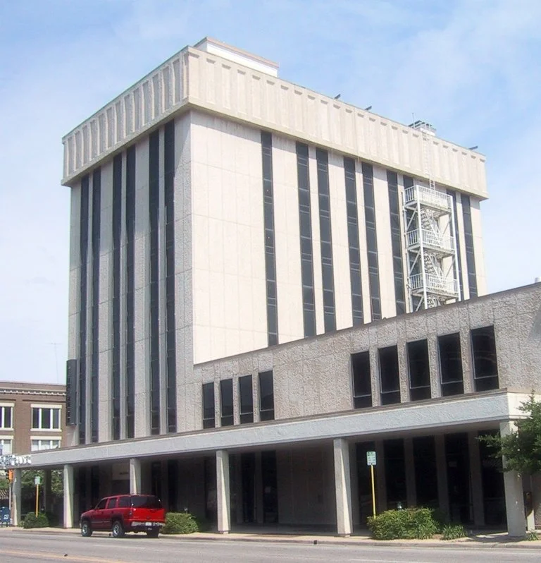A tall, modern beige building with vertical black windows and an external fire escape staircase, with a red vehicle parked underneath on a city street.