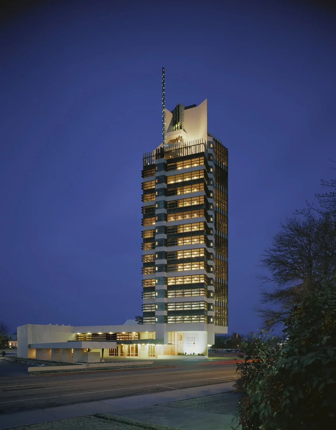 Exterior of Price Tower in Bartlesville, OK with illuminated windows at night, featuring unique architectural design elements and an LED light strip on top, with a clear dark blue sky and some trees in the foreground.