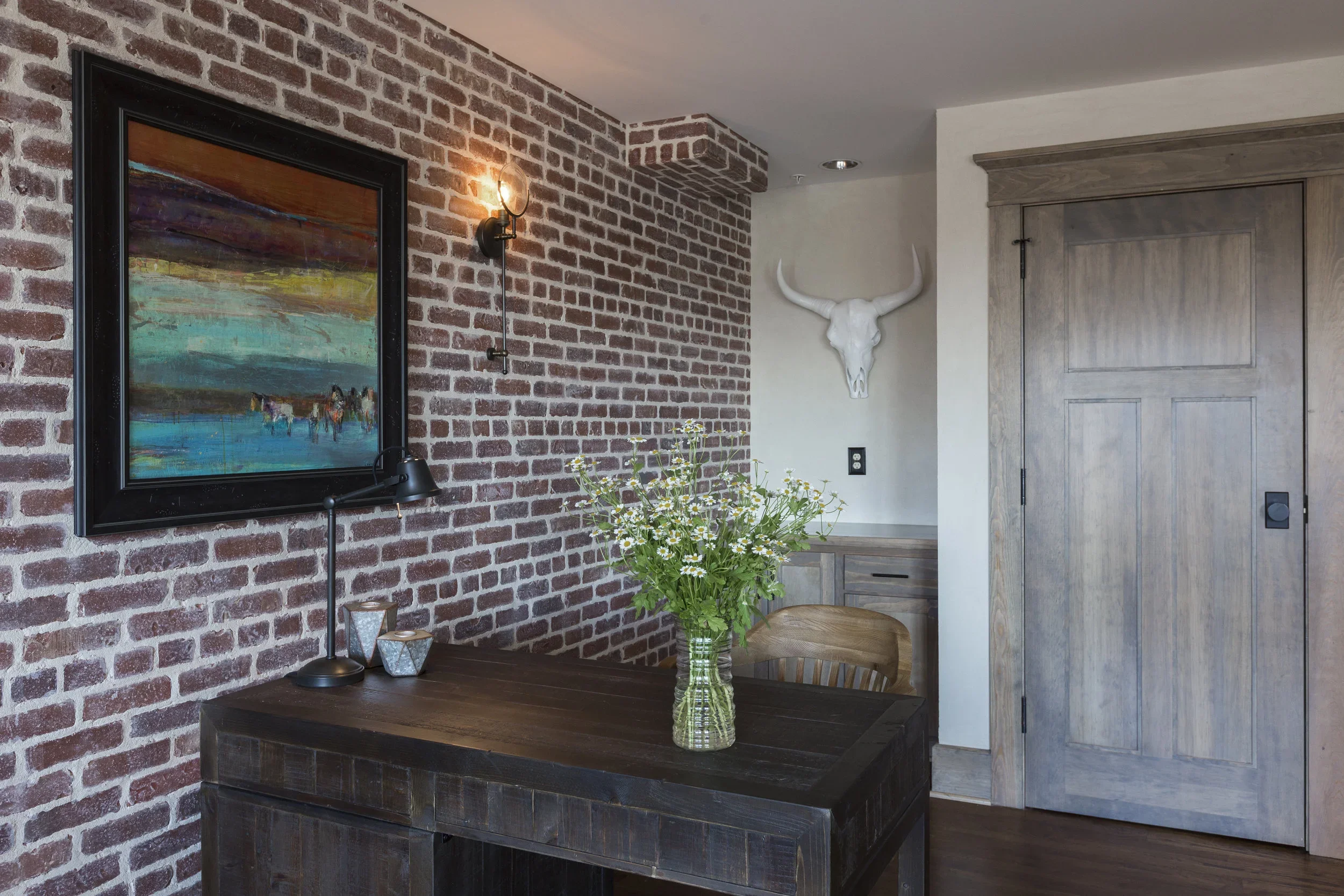 Interior of a room with a brick accent wall, a framed colorful landscape painting, a black desk lamp, a glass vase with white daisies on a dark wood table, a wooden chair, a mounted white animal skull with horns, and a wooden sliding door.