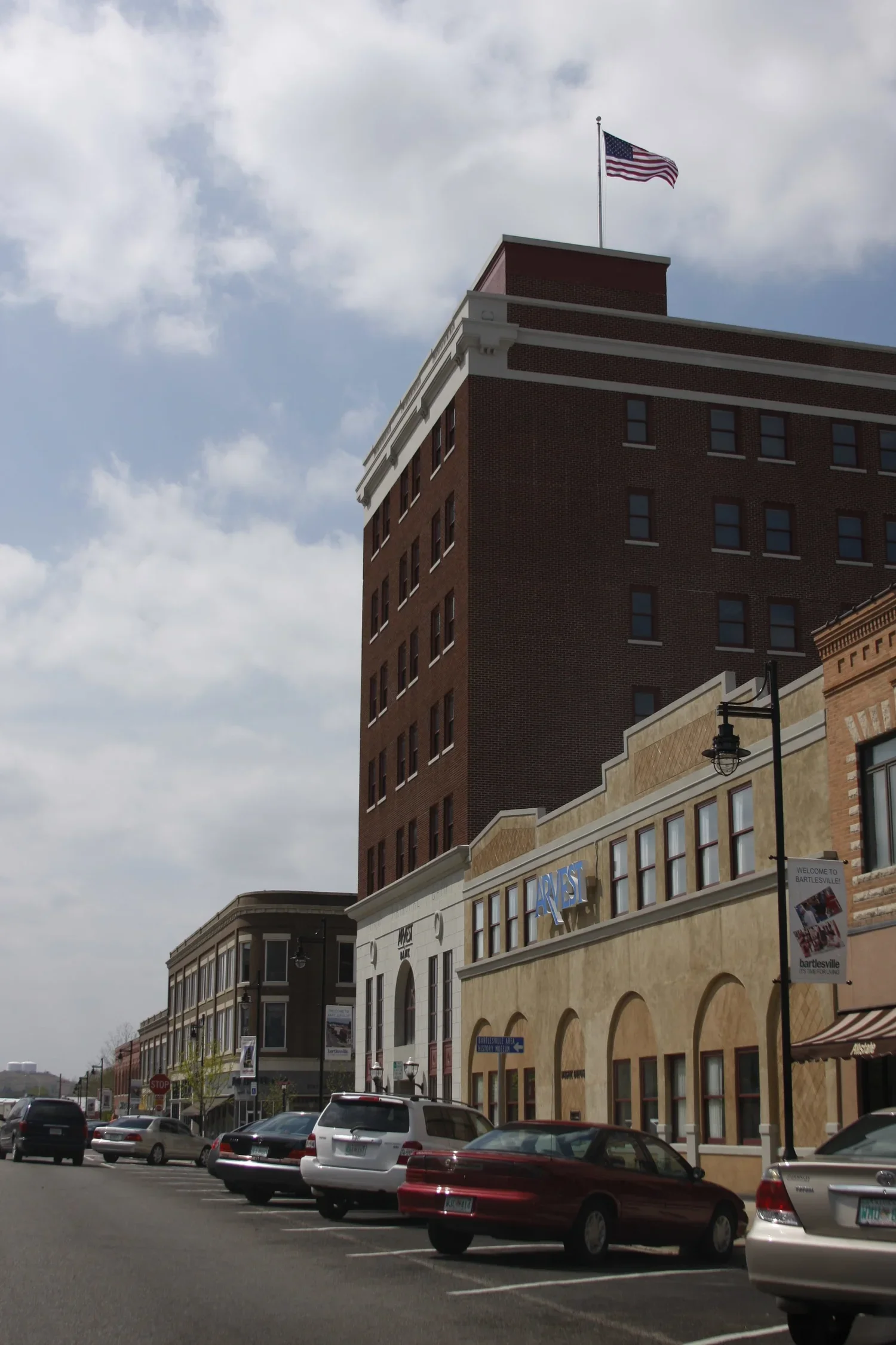 Street view of multiple buildings, including a tall brick building with an American flag on top, parked cars along the sidewalk, and a cloudy sky.