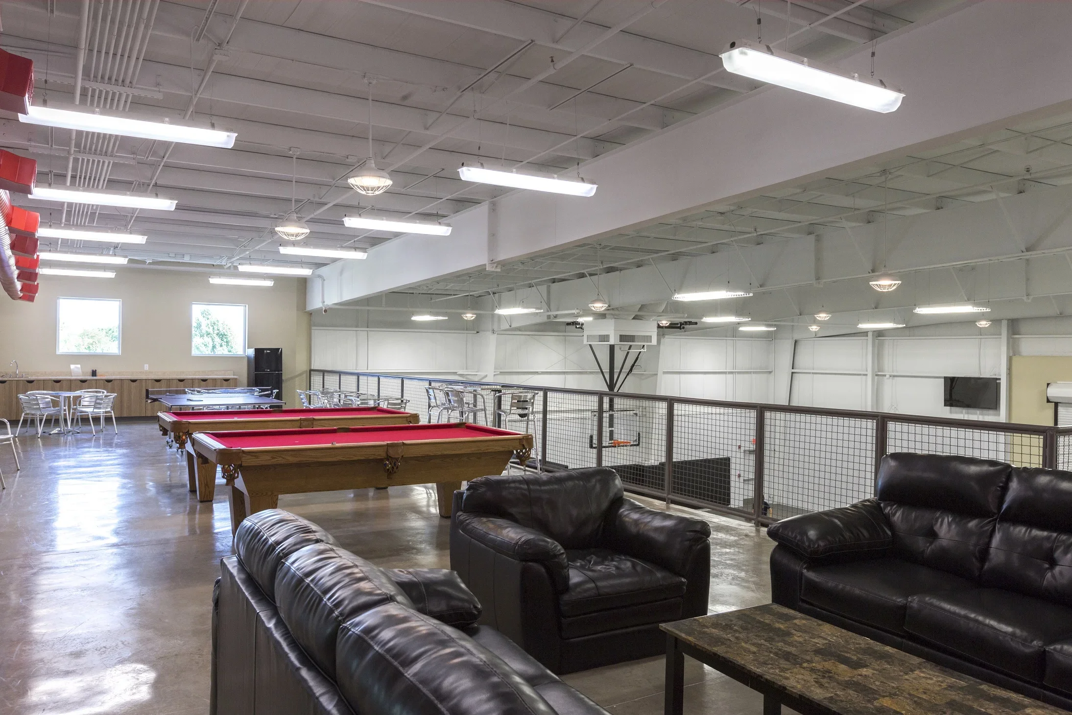 Indoor recreational space with black leather couches, a pool table with a red felt top, ping pong tables, and a balcony with seating. Overhead fluorescent lighting and large windows are visible.