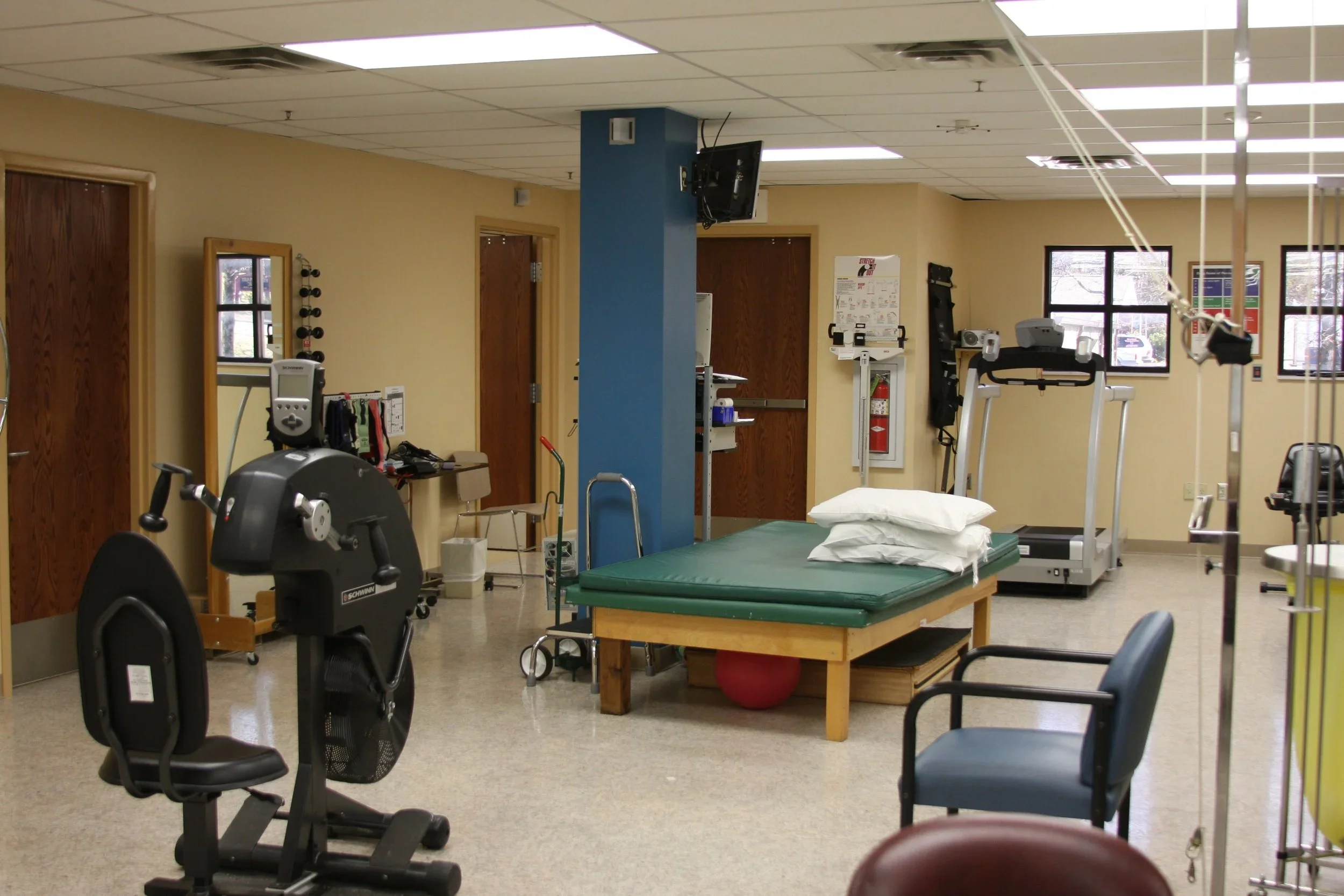 A hospital or medical rehabilitation room with exercise equipment, a bed, and chairs, with several windows and a blue wall.