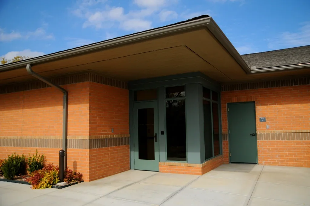 Exterior view of a brick building with a green door and windowed corner, with small landscaped bushes in front and a clear blue sky.