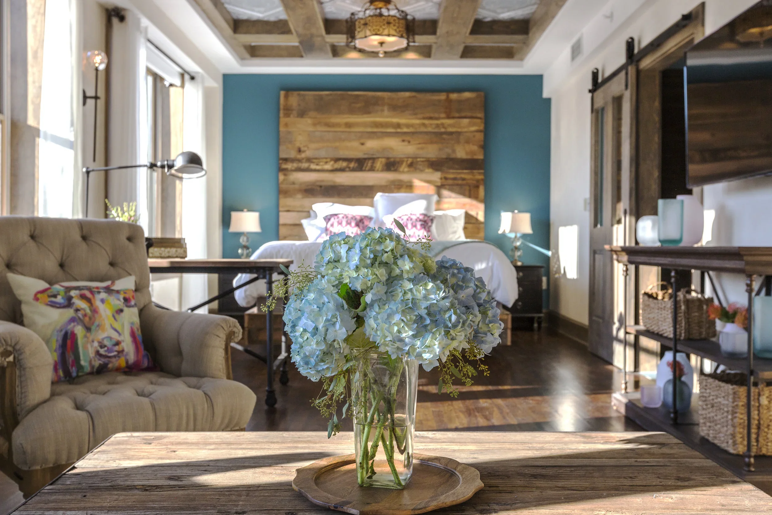 A bedroom with rustic decor, featuring a wooden headboard, blue accent wall, and floral bedding. A vase with light blue flowers is on a wooden table in the foreground, with a beige armchair and a decorative pillow to the left and a shelf with baskets to the right.